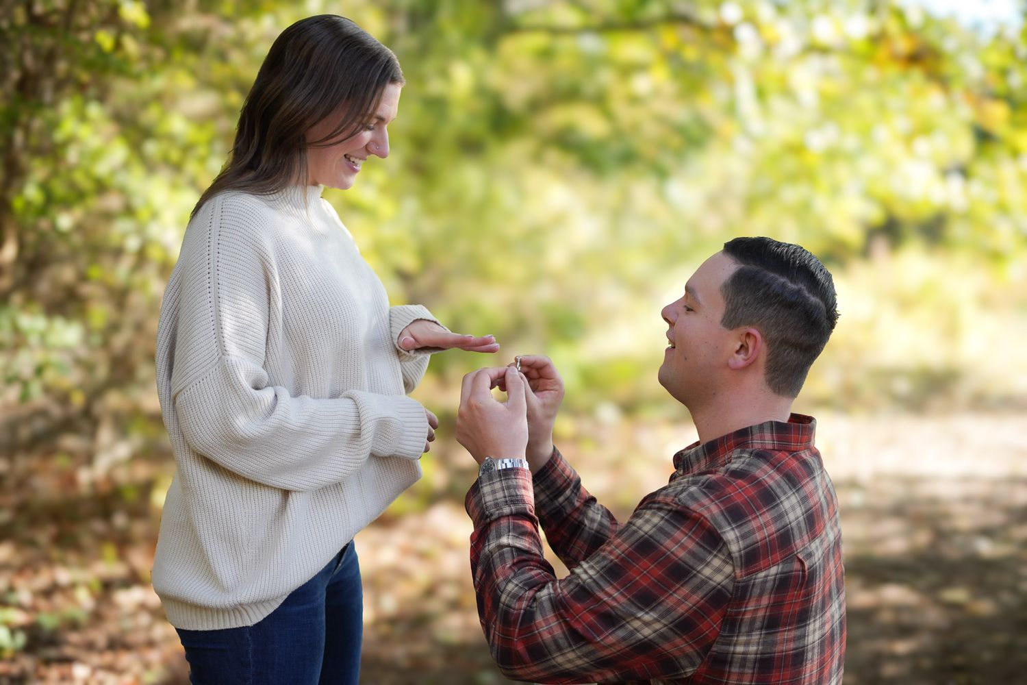 Man on one knee proposing to woman outdoors in autumn forest setting.