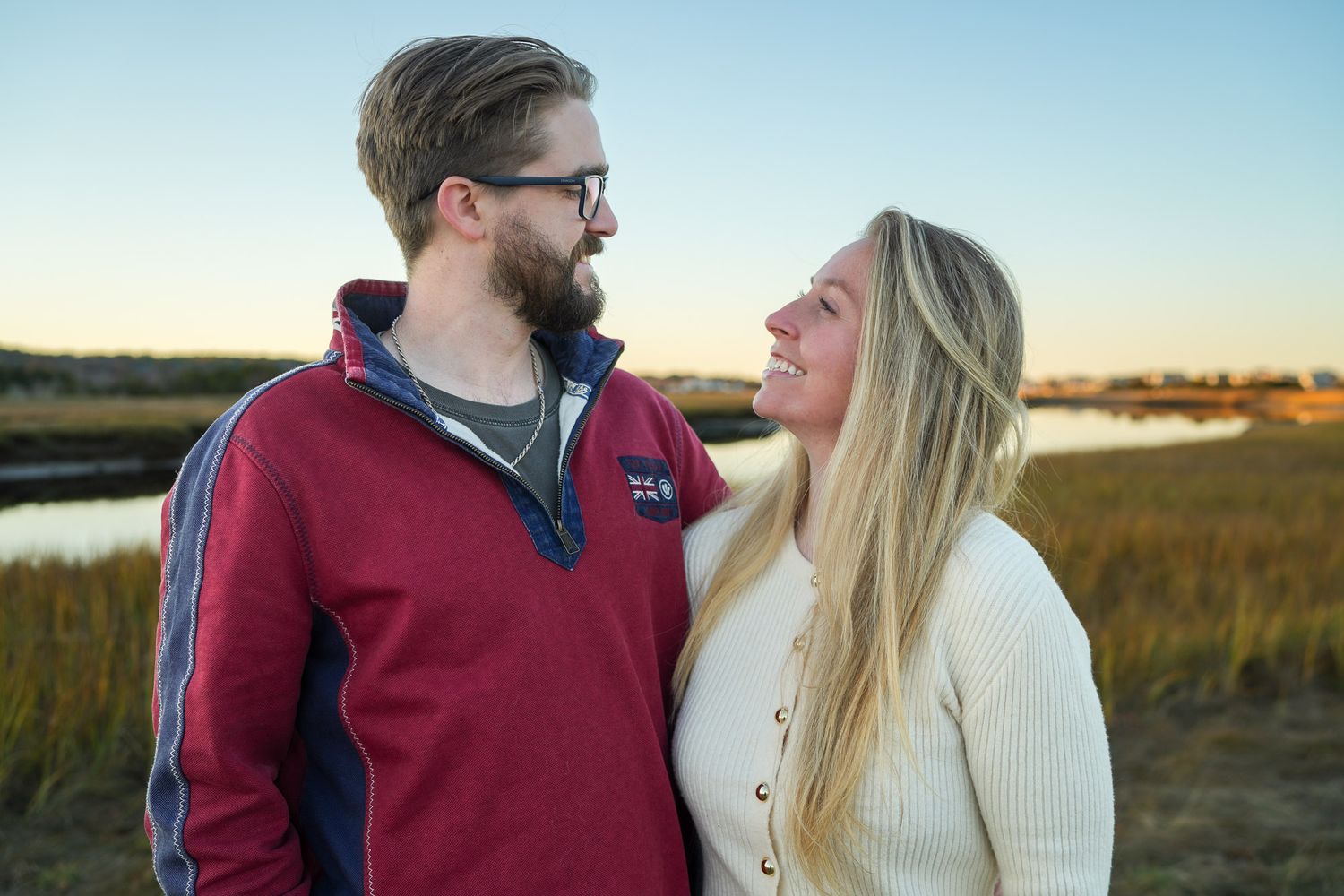 Couple gazing at each other near a marsh at sunset, man in red pullover and woman in cream sweater.