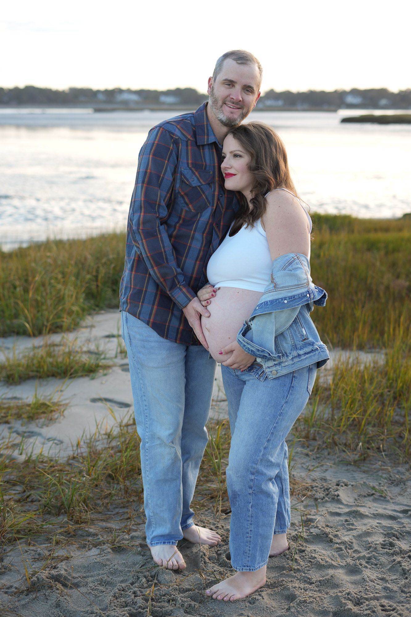 Man embracing pregnant woman from behind as they pose together on sandy beach at sunset.