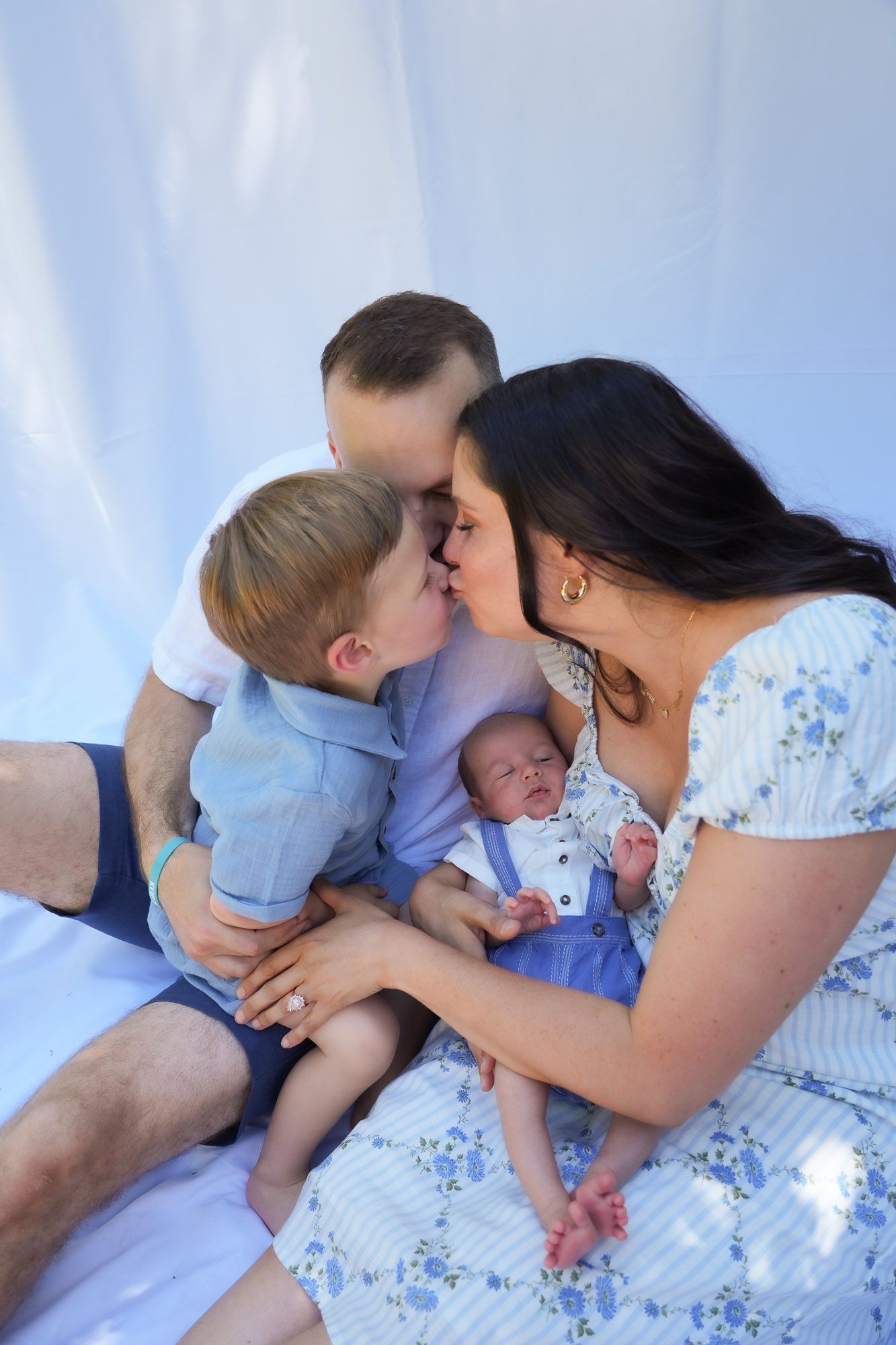 Family of four sharing a kiss with toddler and newborn on white backdrop.