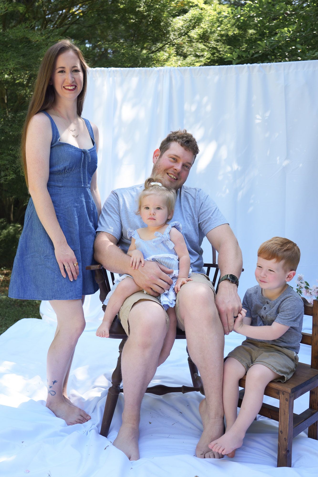 Smiling family of four enjoys outdoor photo session with white sheet backdrop in sunny backyard setting.