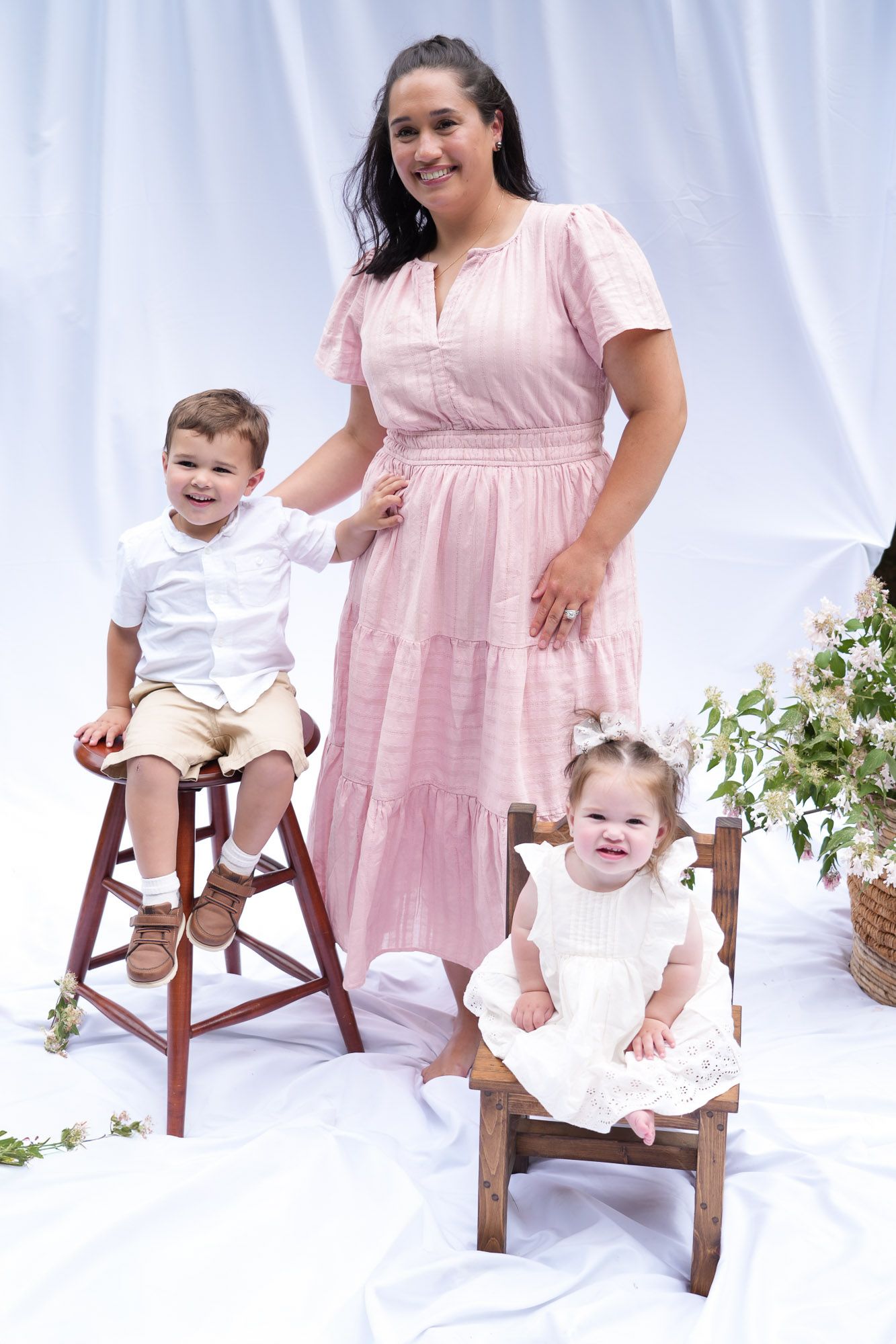 Smiling woman in pink tiered dress stands outdoors with two young children seated on wooden chairs against white backdrop.