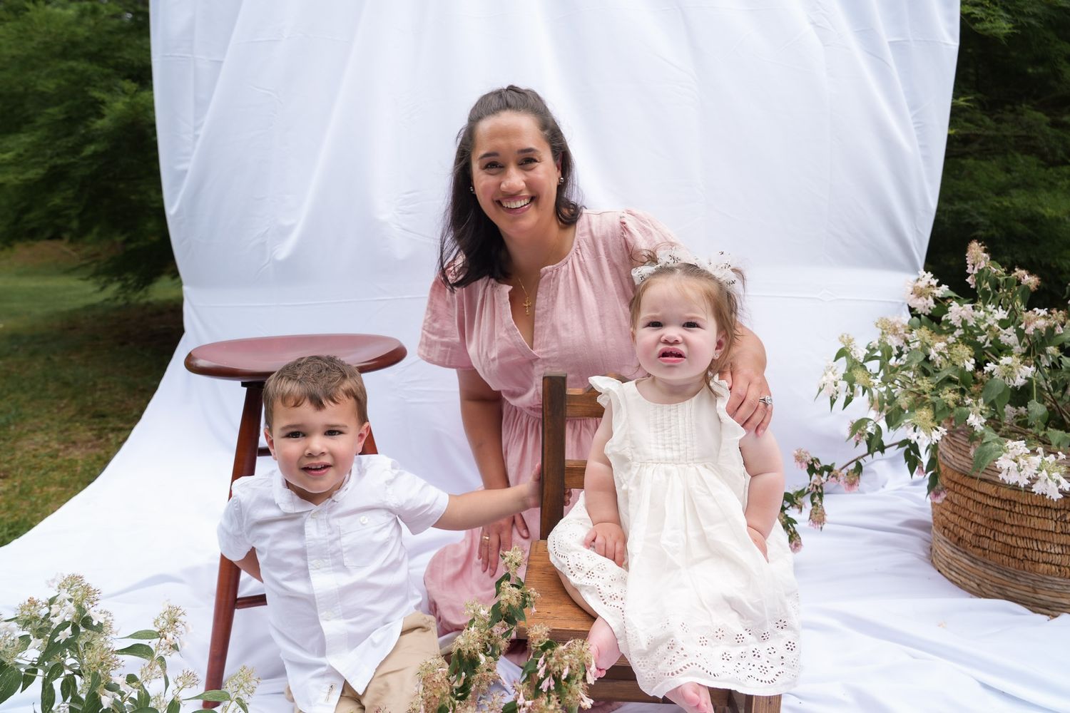 Mother in pink dress posing with young boy and baby girl in white dress at outdoor photo session.
