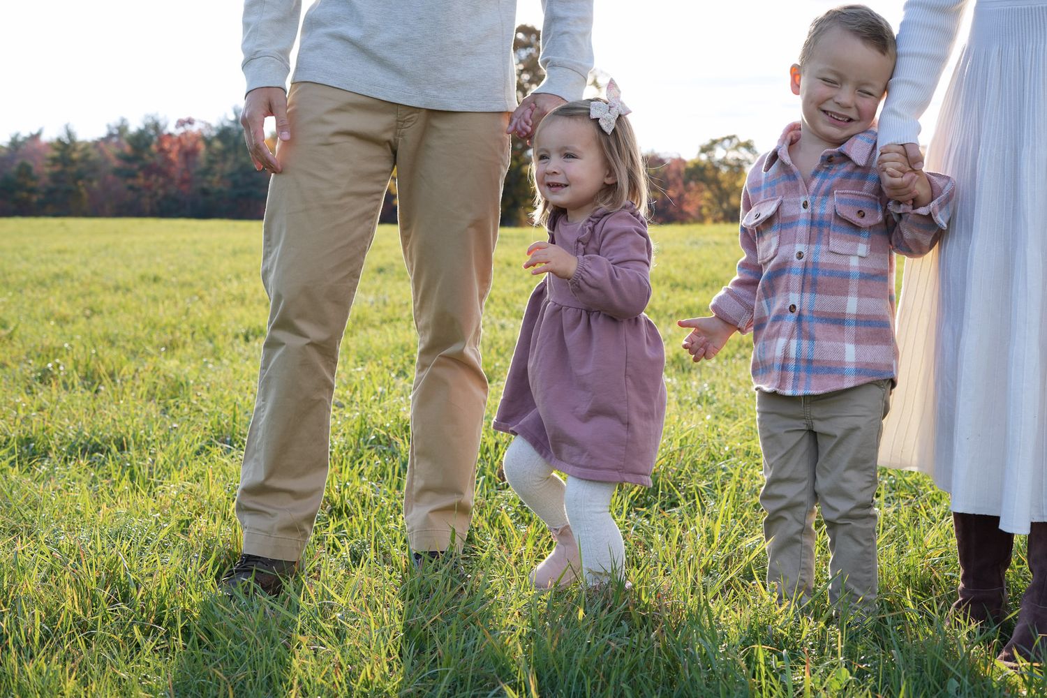 Two young children holding parents hands while walking through a sunny green field in autumn.
