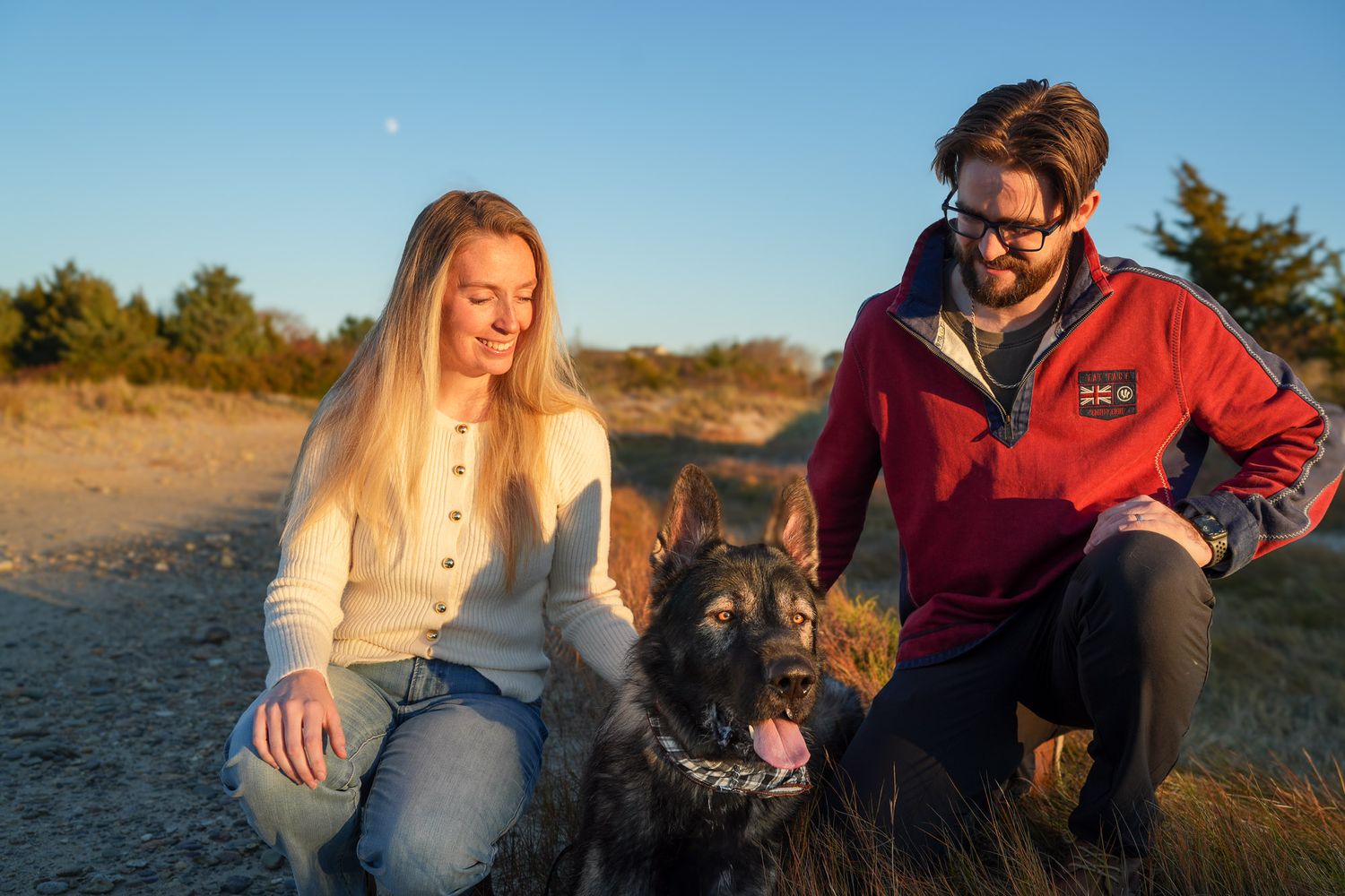 Couple crouching with their German Shepherd dog on a sandy beach path at sunset.