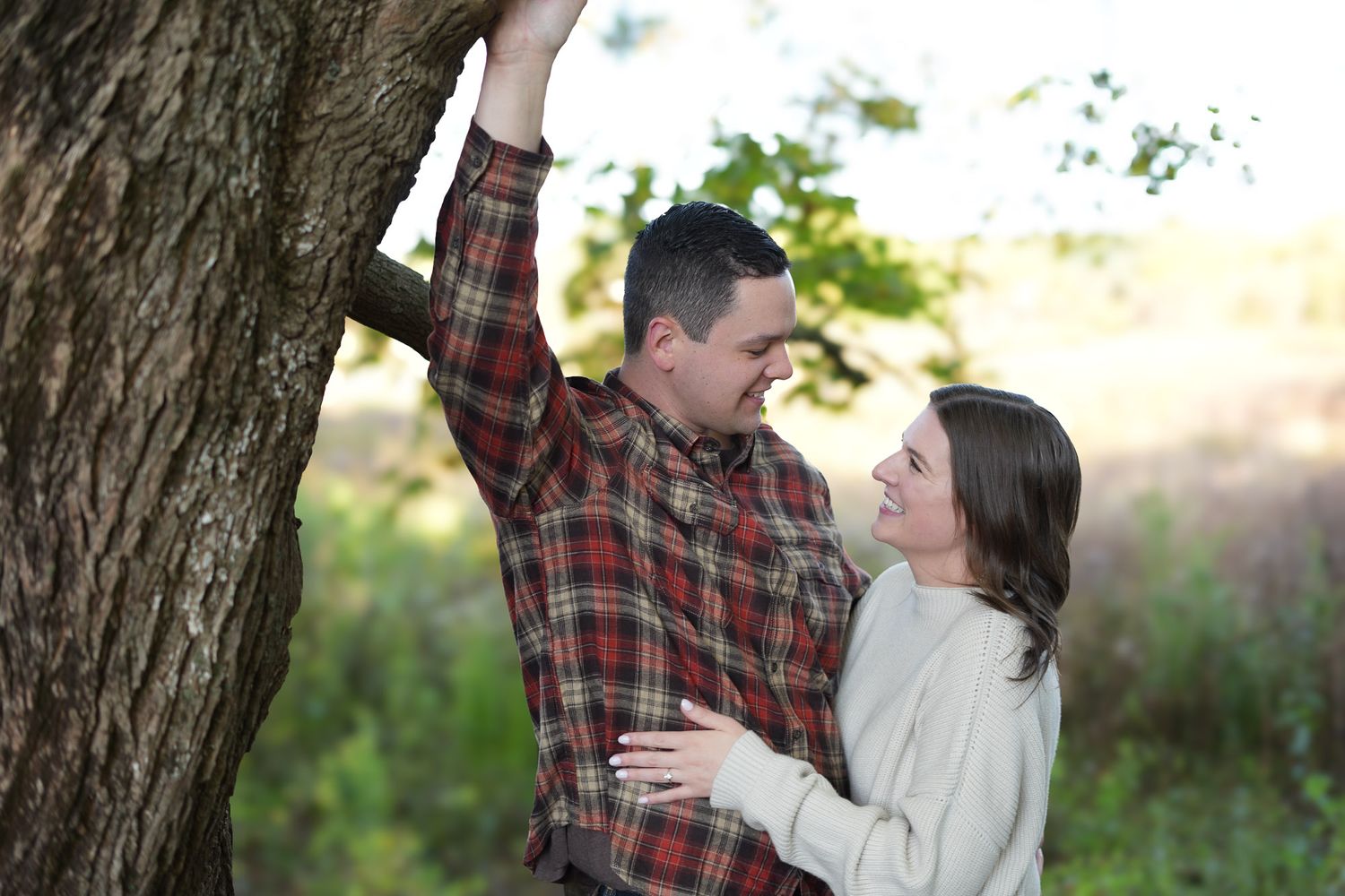 Couple sharing a romantic moment near a tree trunk in a sunlit outdoor setting.