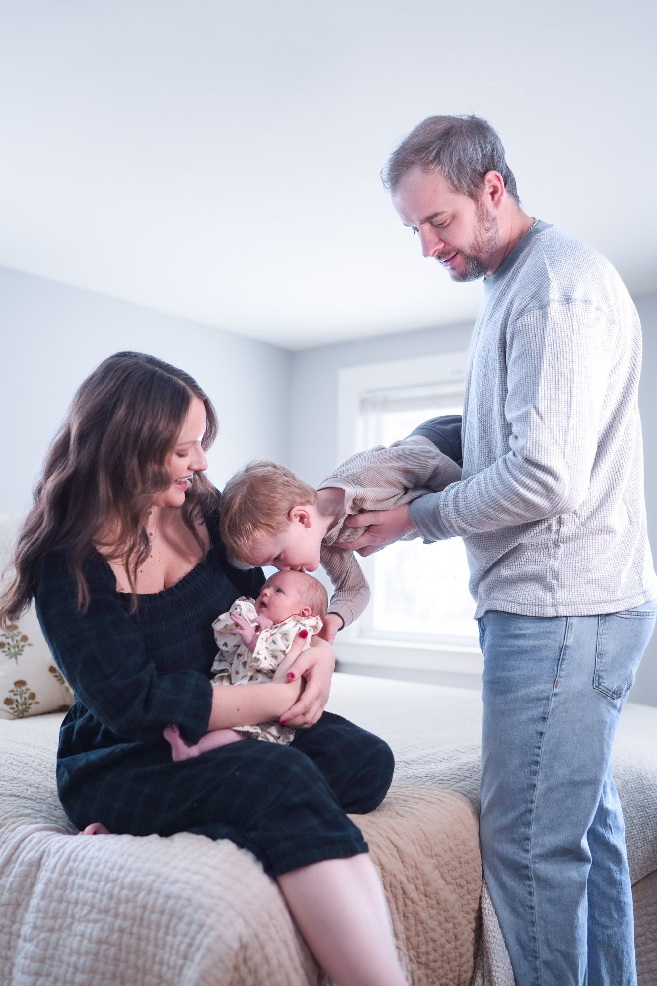Family with mother holding newborn baby as toddler leans in to kiss while father watches.