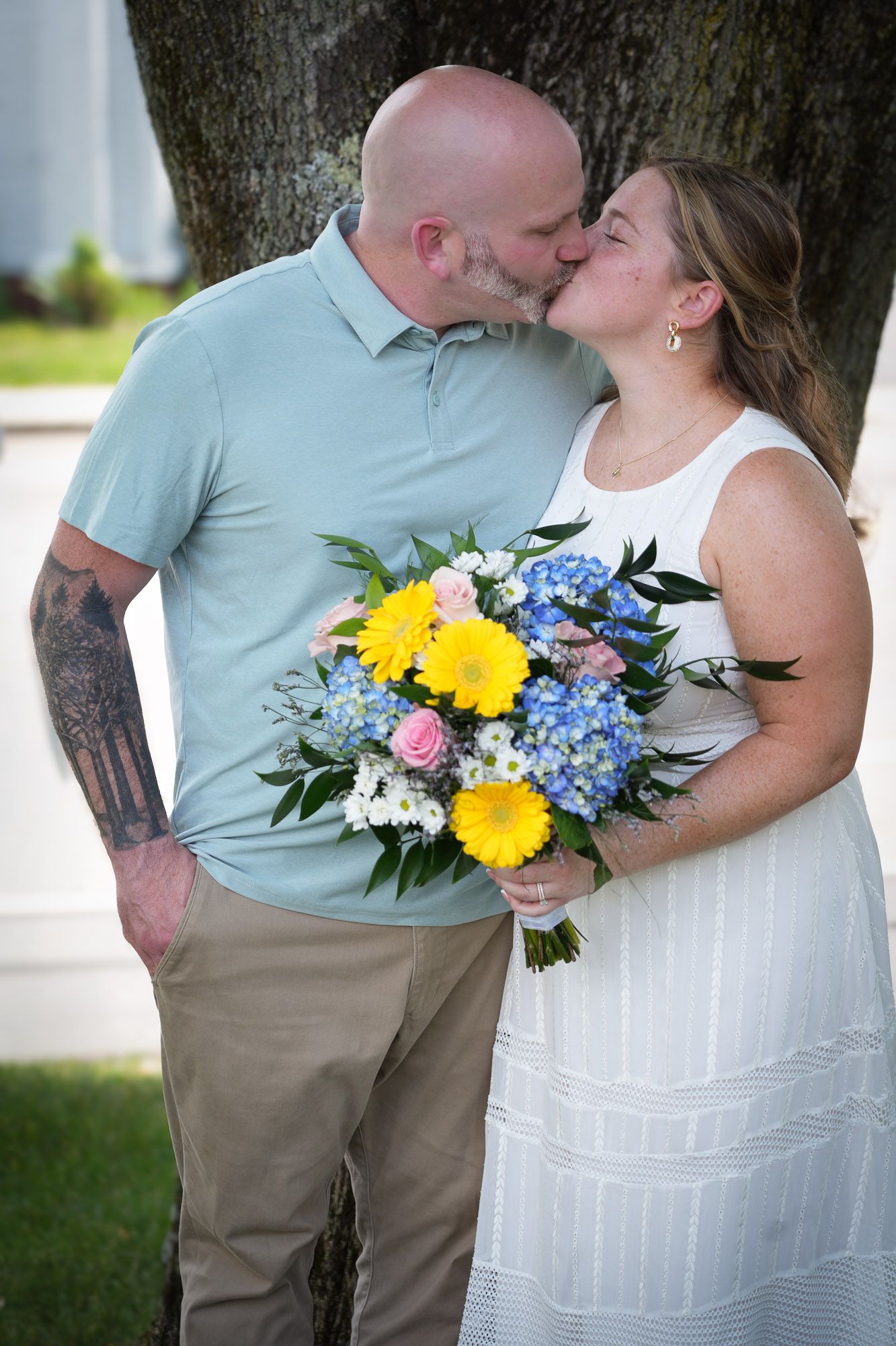 A couple shares a kiss while the bride holds a colorful bouquet of yellow and blue flowers.