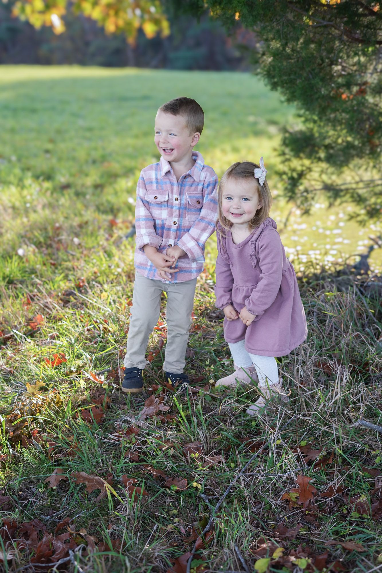 Two young siblings laughing and smiling together outdoors among fallen autumn leaves in a park.