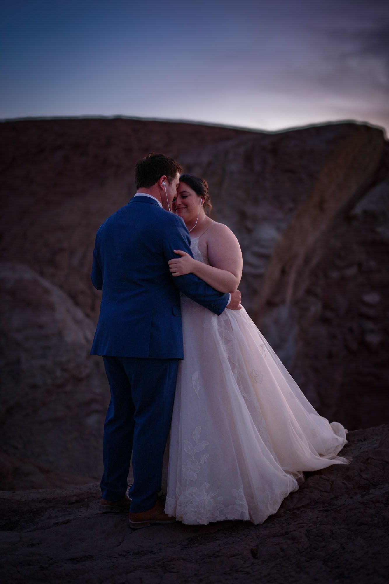 Couple in wedding attire share a romantic embrace at dusk against dramatic rocky desert backdrop in Death Valley.