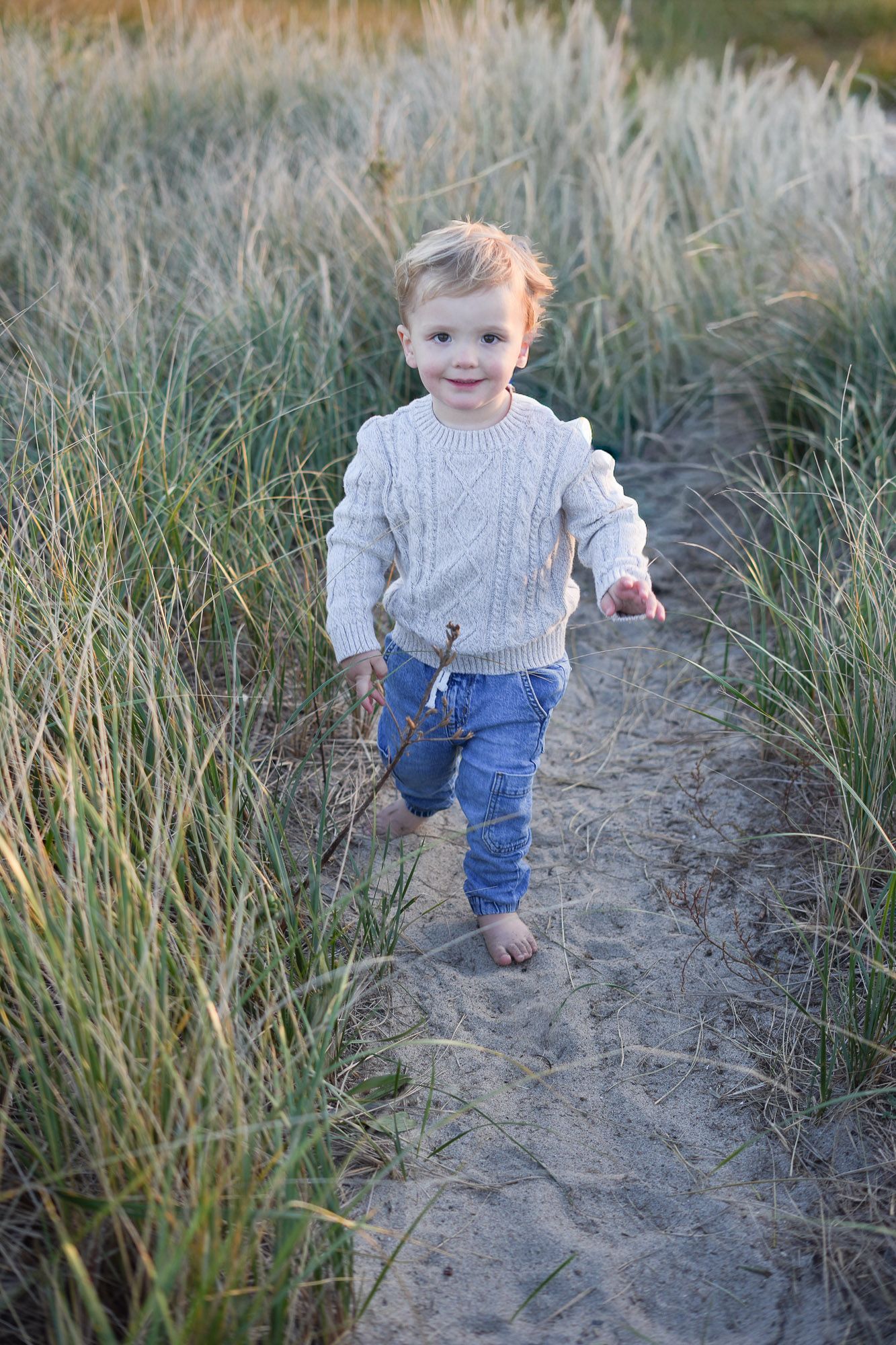 Happy toddler boy in cream sweater running barefoot through tall beach grass.