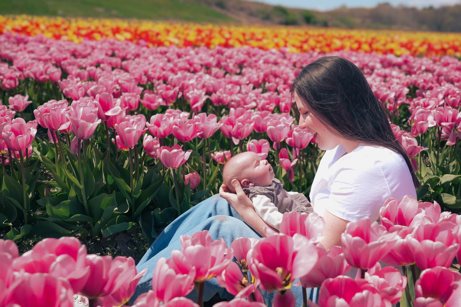Woman sitting in a vibrant pink tulip field, tenderly holding a newborn baby in spring.