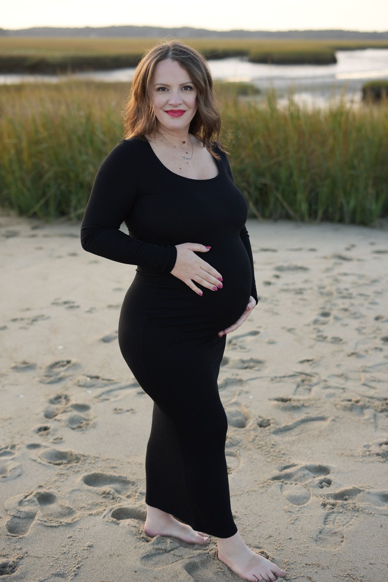 Pregnant woman in black maxi dress holding belly on sandy beach at sunset.