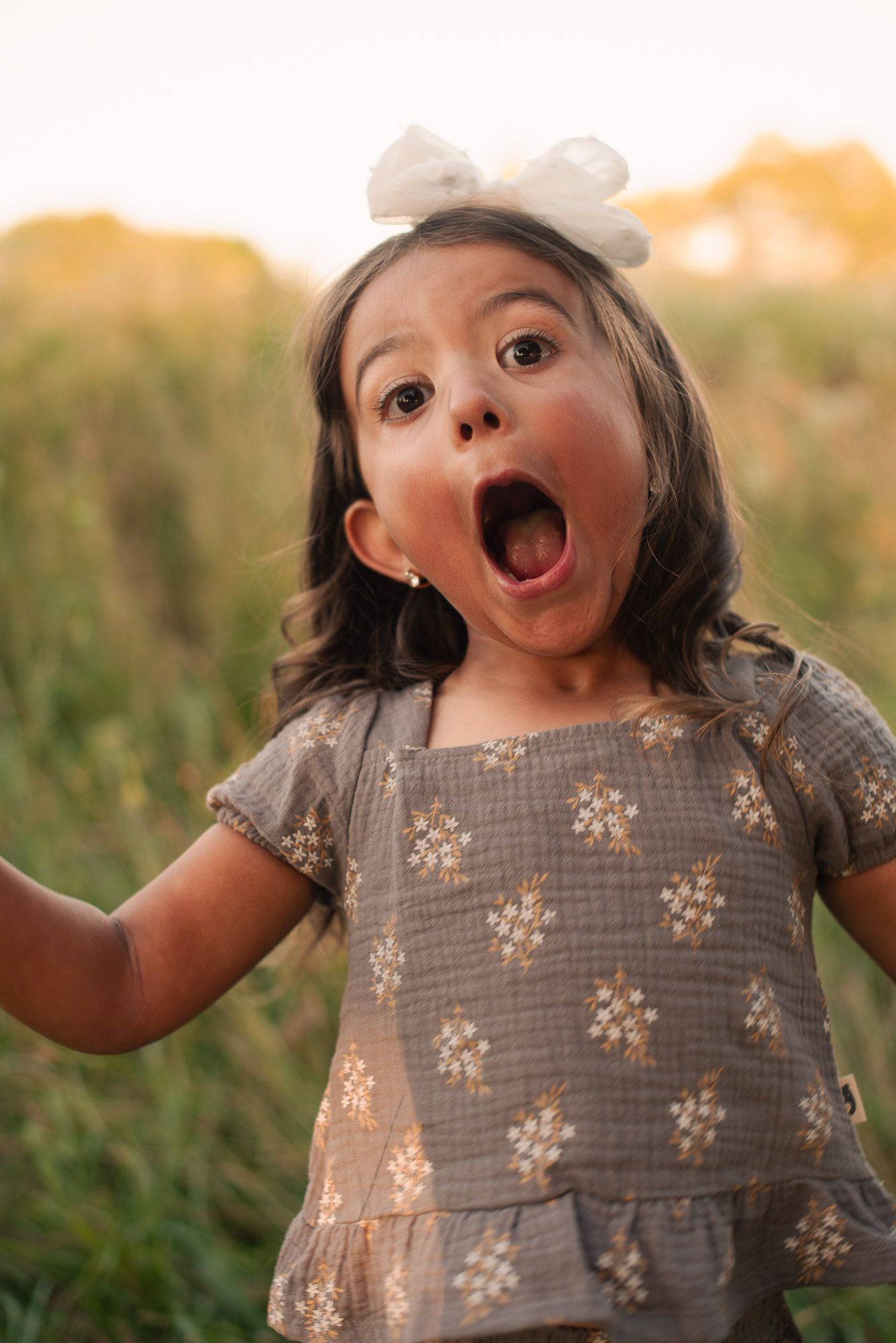 A surprised toddler girl with a white bow headband and floral dress makes a funny face in a sunny field.