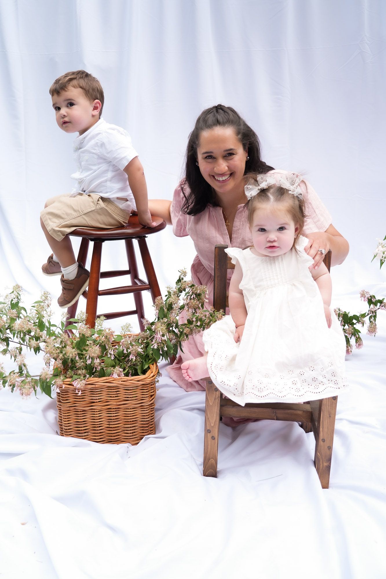 Mother in pink dress posing with toddler boy on stool and baby girl in white dress indoors.