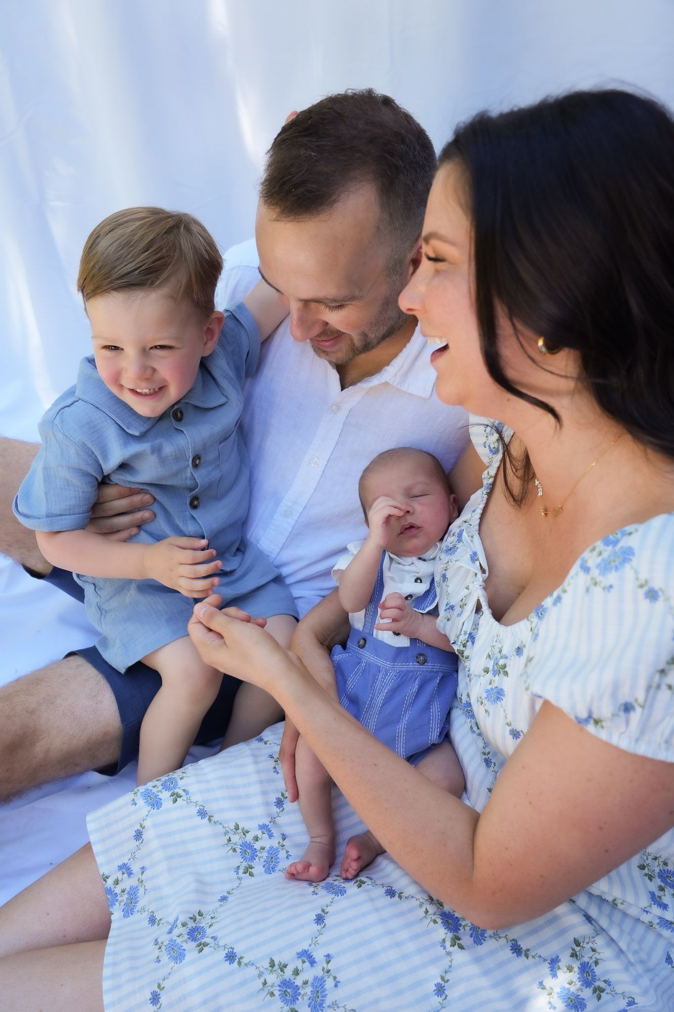 Joyful family of four with toddler and newborn baby sitting together on white fabric.