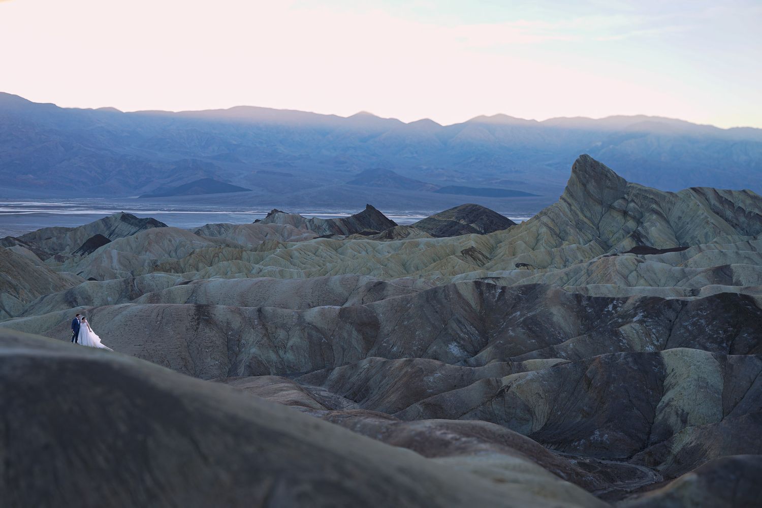 Bride and groom stand tiny against dramatic Death Valley badlands at Zabriskie Point during golden hour.