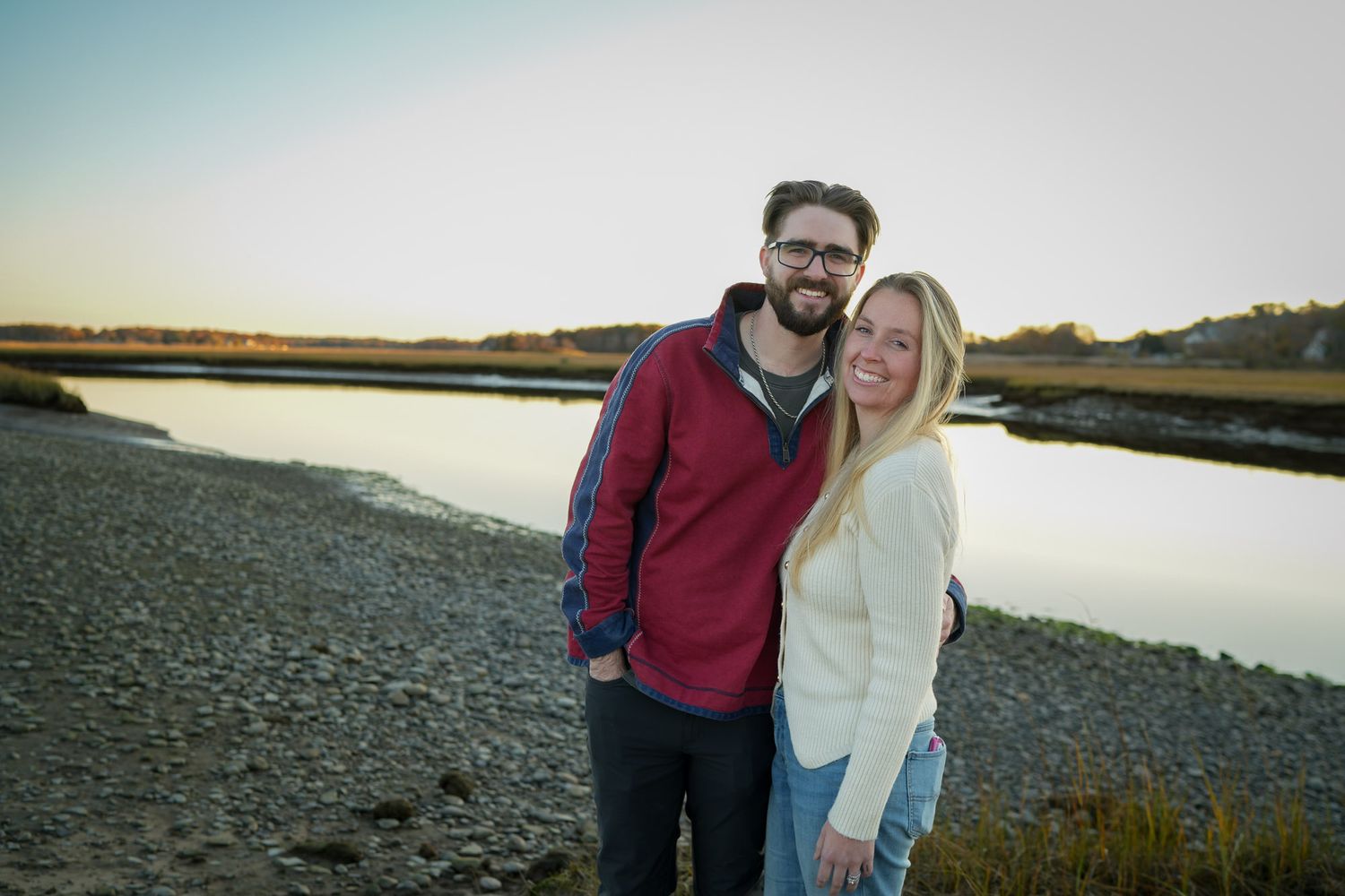 Happy couple embracing on a rocky shoreline near a calm marsh at golden hour.