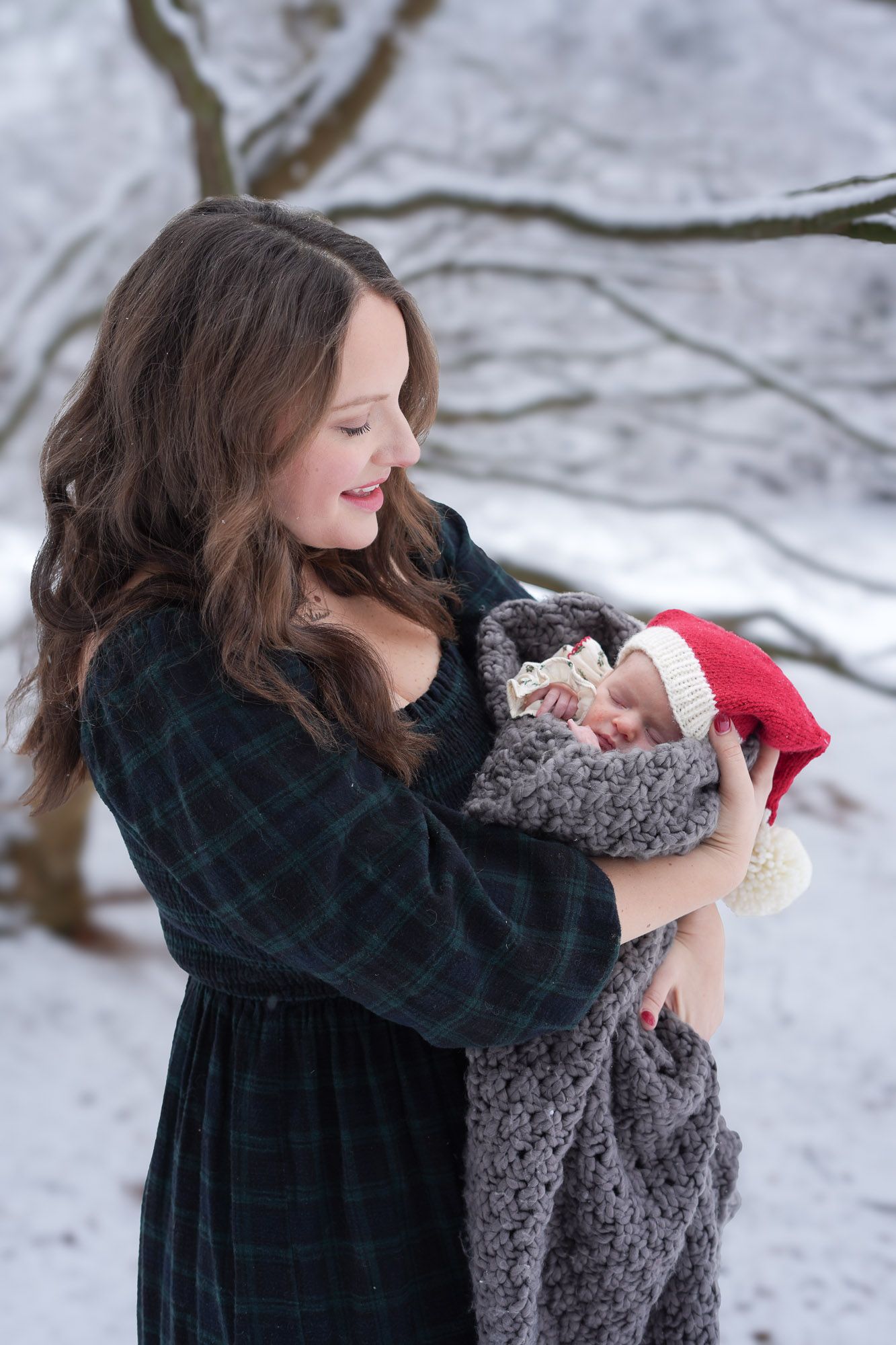 New mother gazing lovingly at swaddled newborn in Santa hat during snowy outdoor winter photo.