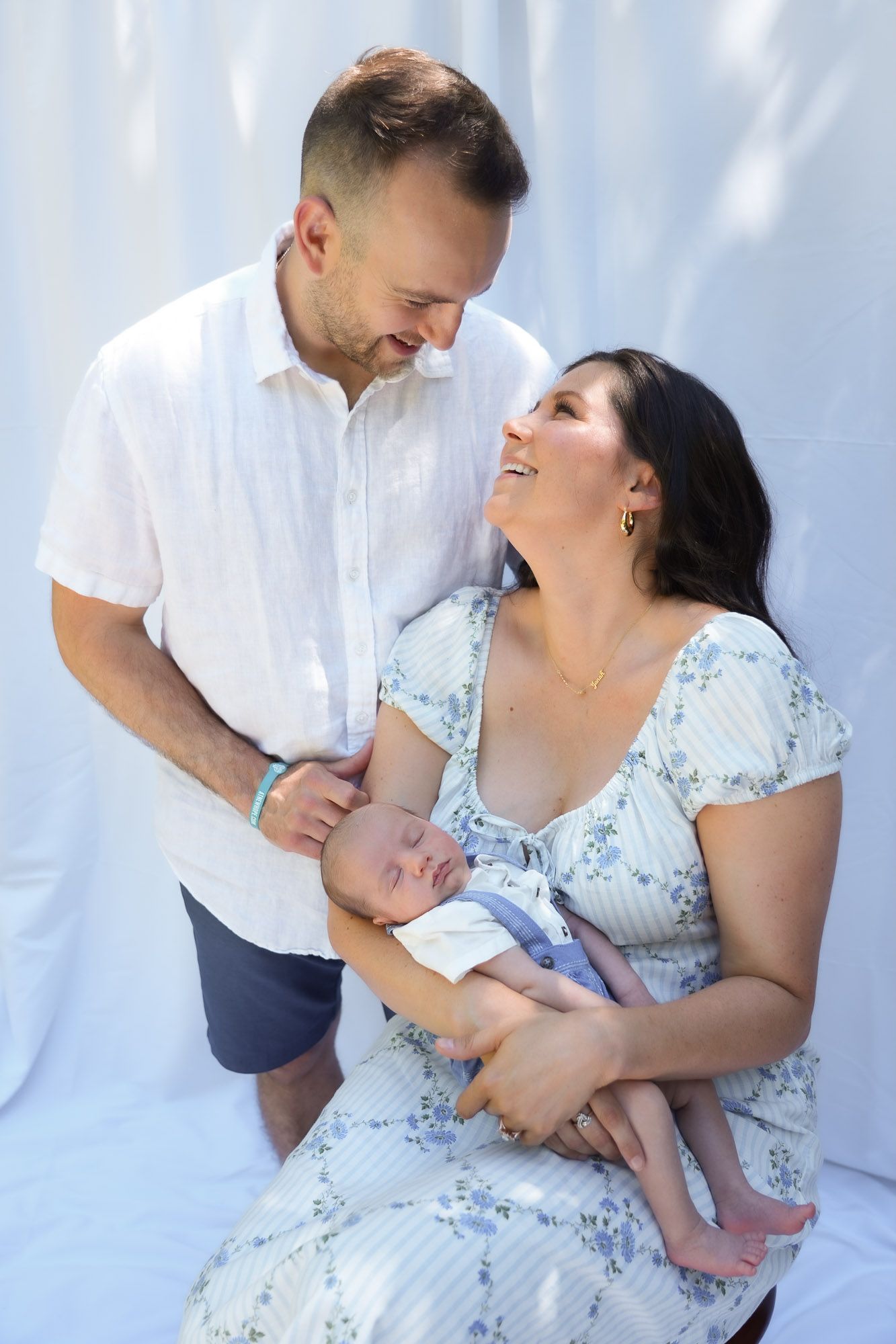 Smiling couple with newborn baby posing together against white backdrop outdoors.
