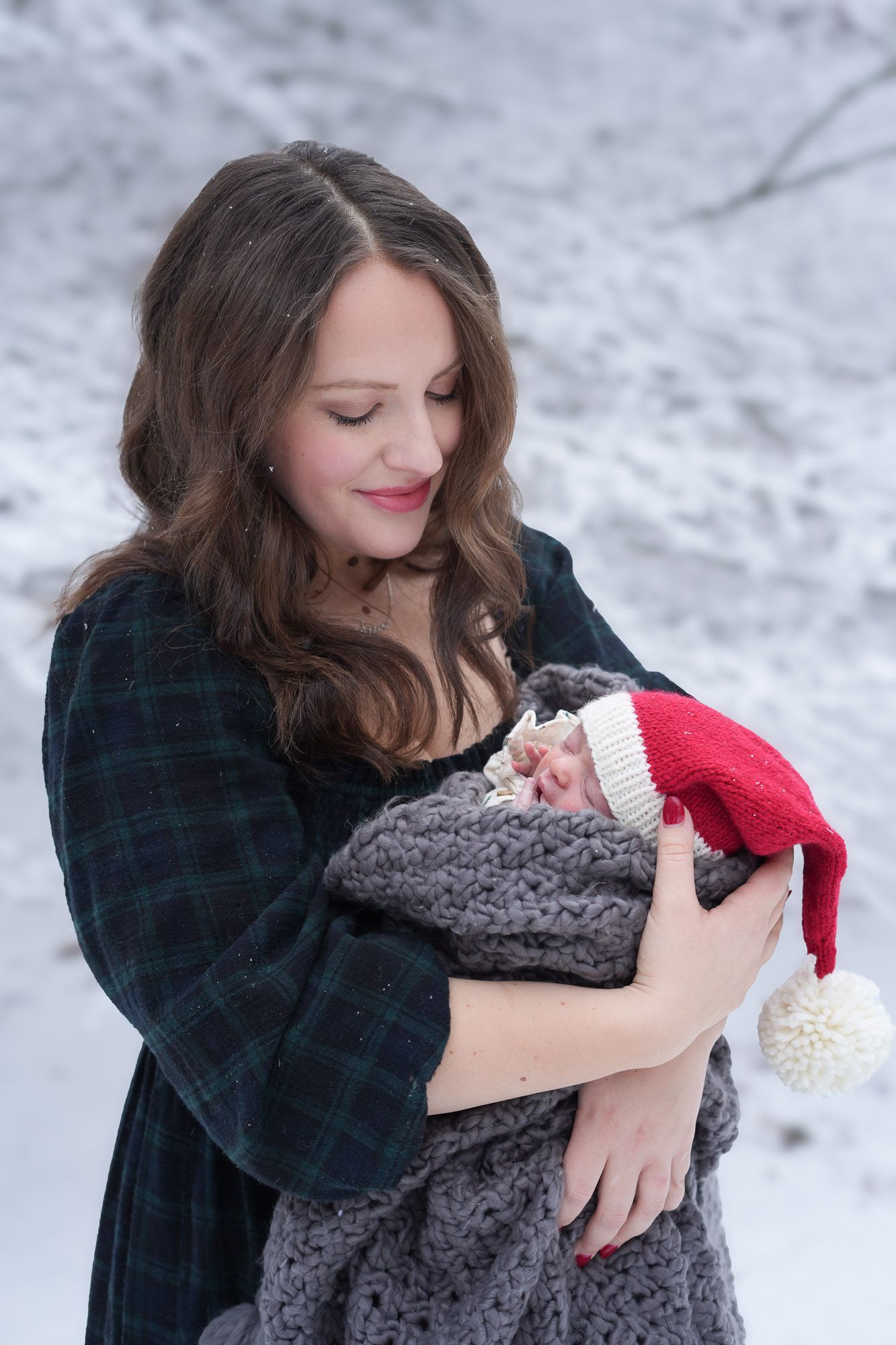 Mother holding newborn baby wrapped in gray knit blanket wearing Santa hat in snowy winter wonderland.