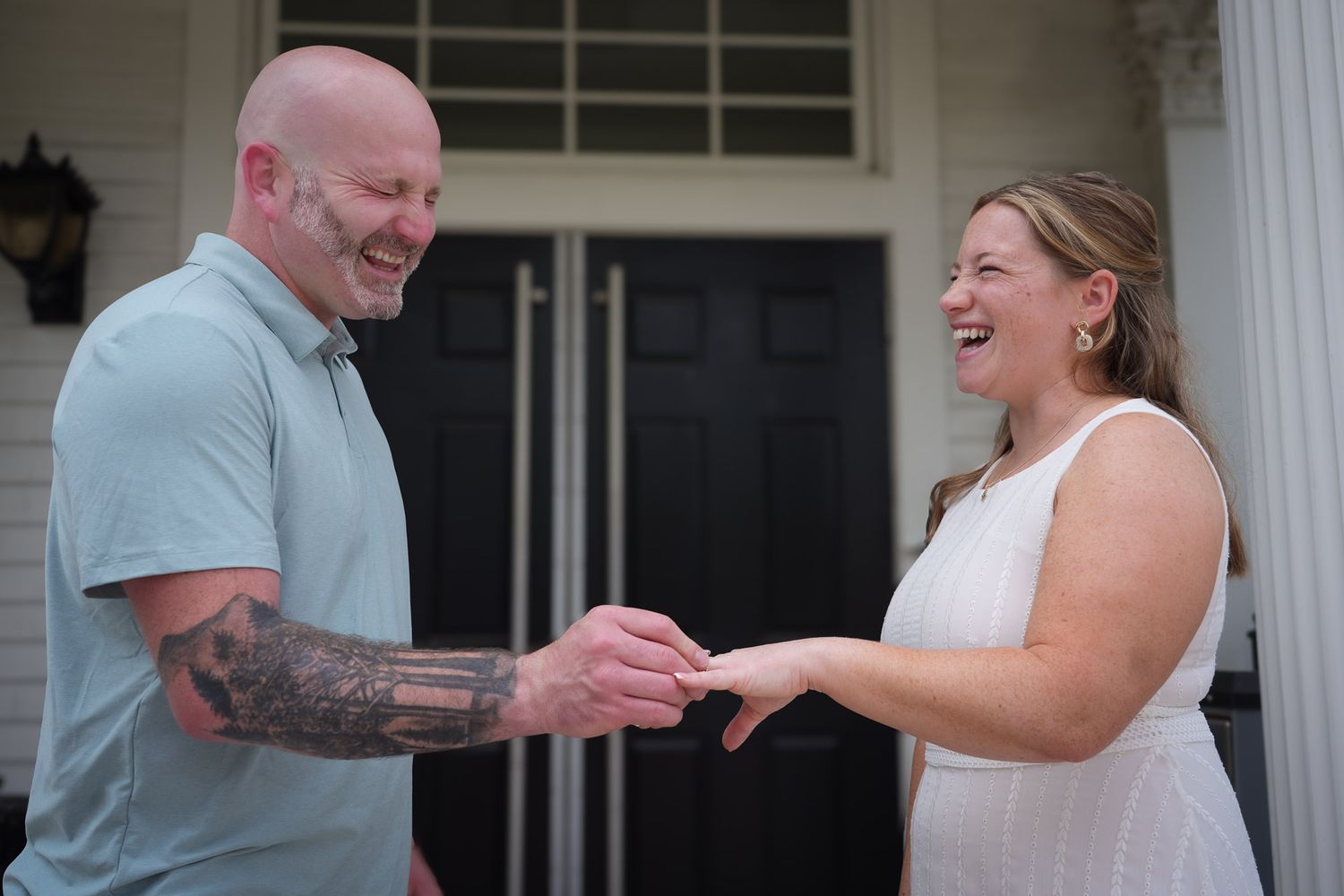 Couple exchanging wedding rings on porch, groom with tattooed arm placing ring on laughing bride's finger.