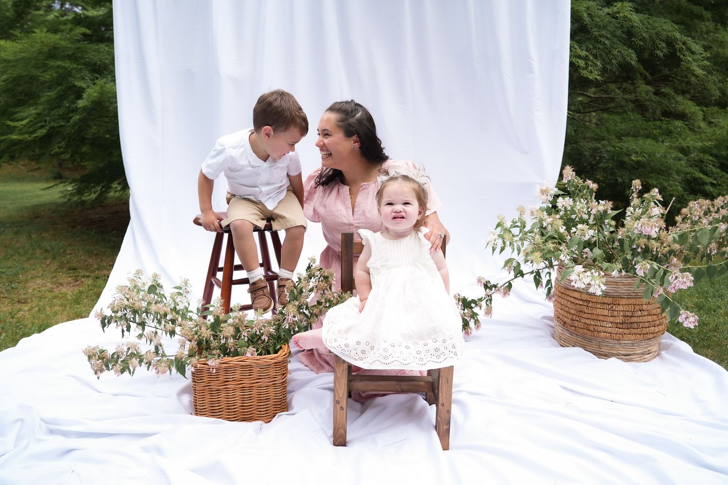 Mother in pink dress with toddler boy on stool and baby girl in white dress at outdoor photo session.