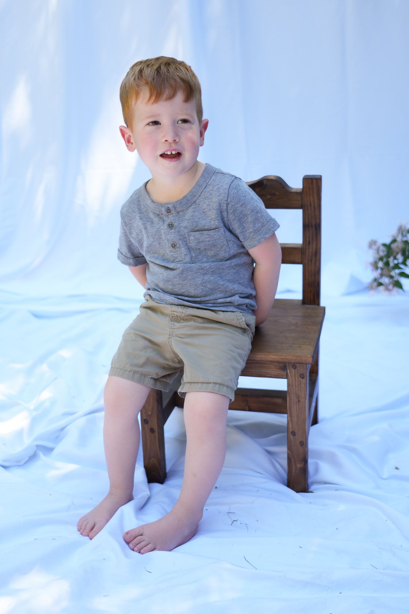 Redhead boy in gray henley shirt and khaki shorts posing on a rustic wooden chair.