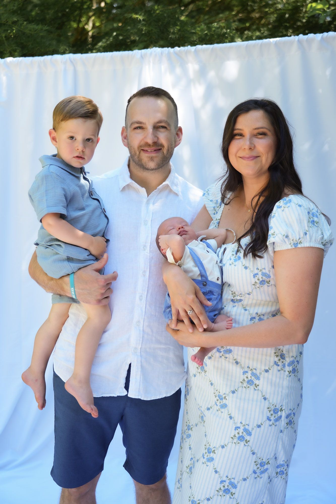 Parents standing outdoors holding toddler and newborn baby in blue outfits.