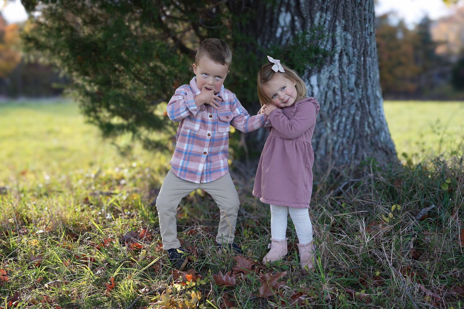Two young children pose playfully near a large tree in an autumn park setting.