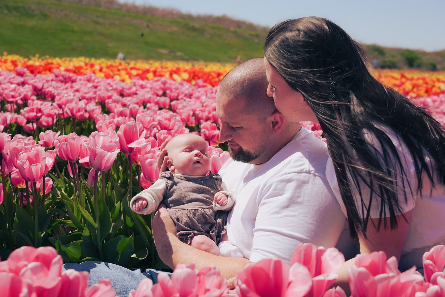 Two parents lovingly look at their newborn baby while sitting surrounded by colorful pink tulips.