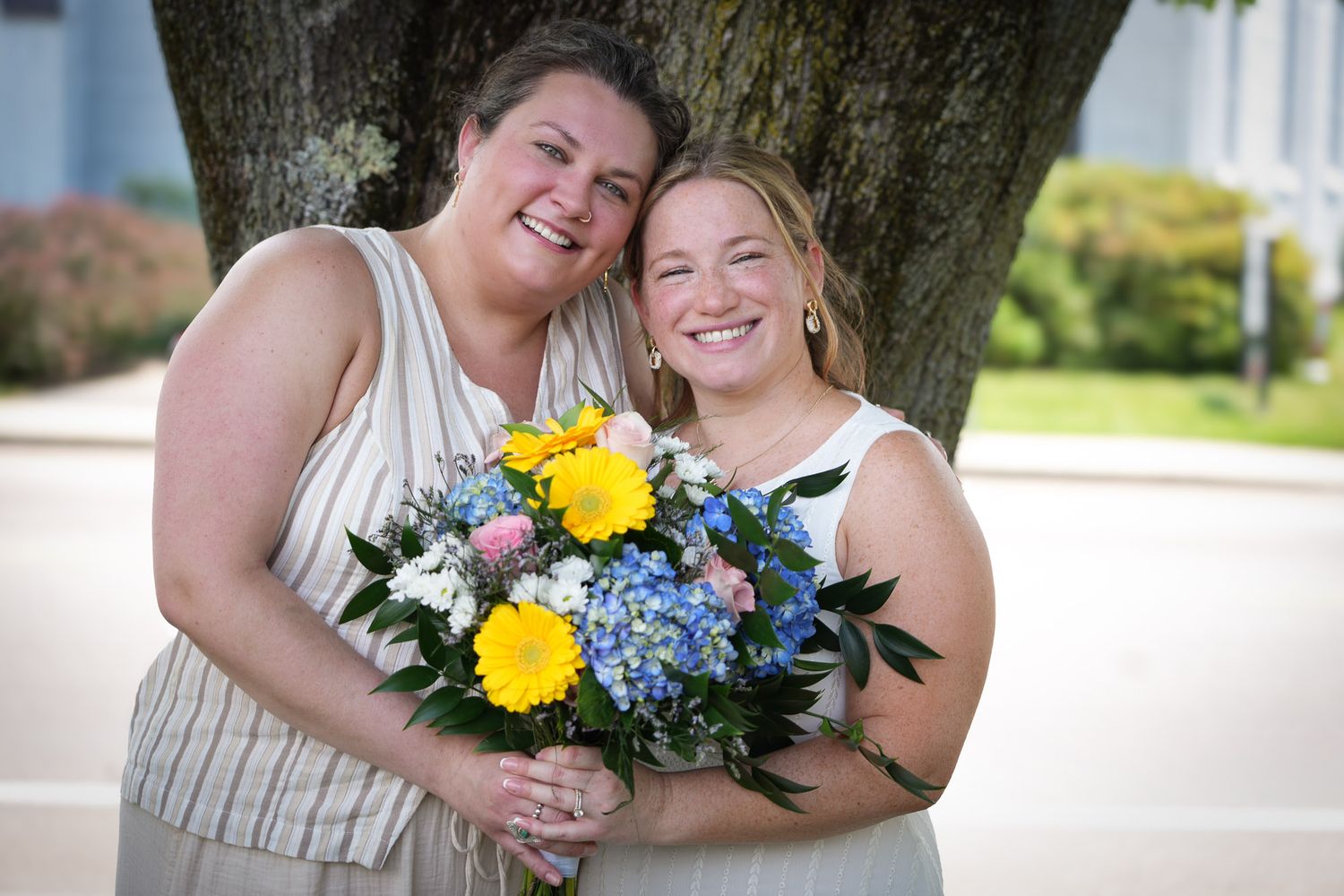Two smiling women pose together outdoors, one holding a vibrant bouquet of yellow and blue flowers.