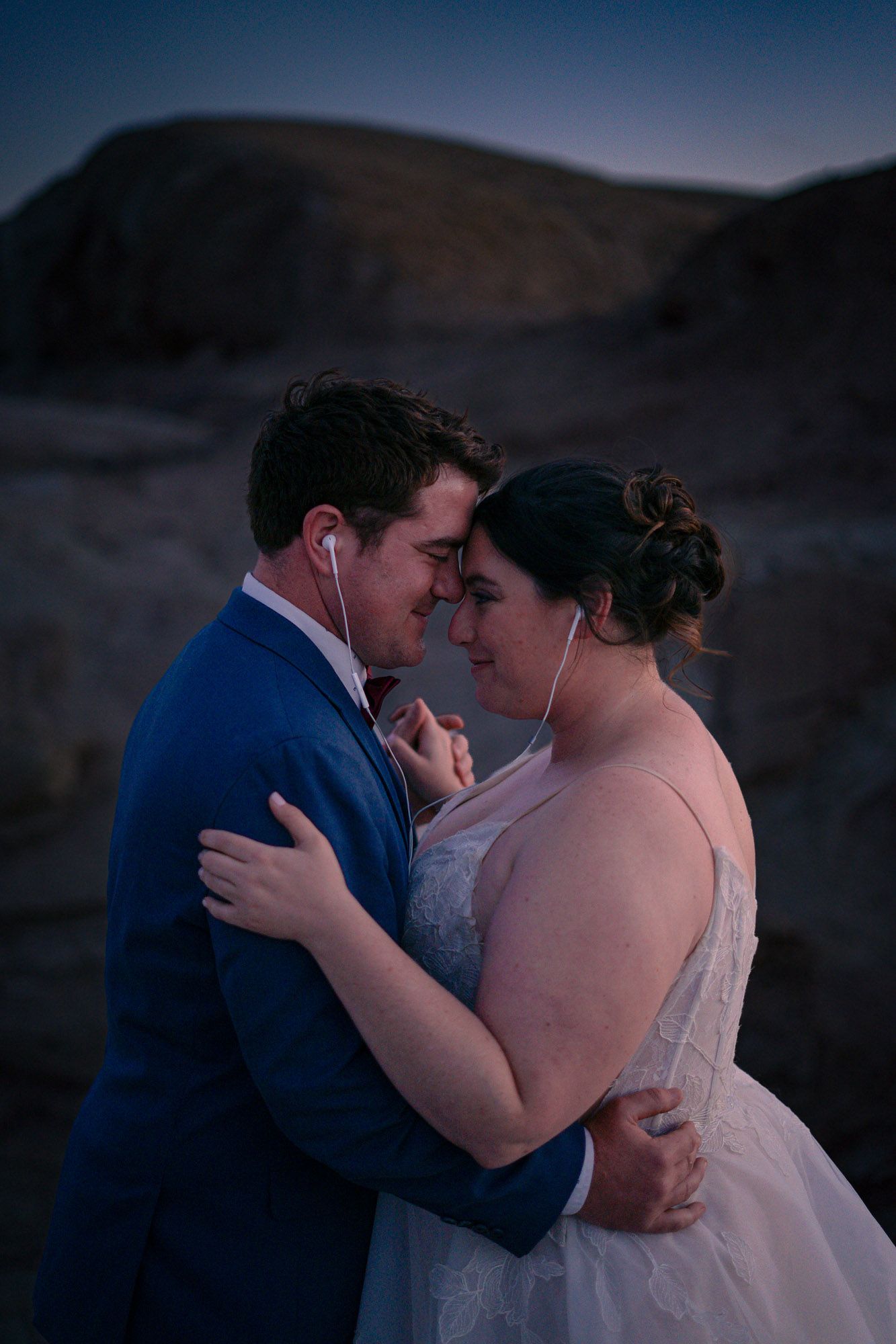 Newlyweds slow dance sharing earbuds in an intimate moment at blue hour against a rugged desert rock formation.