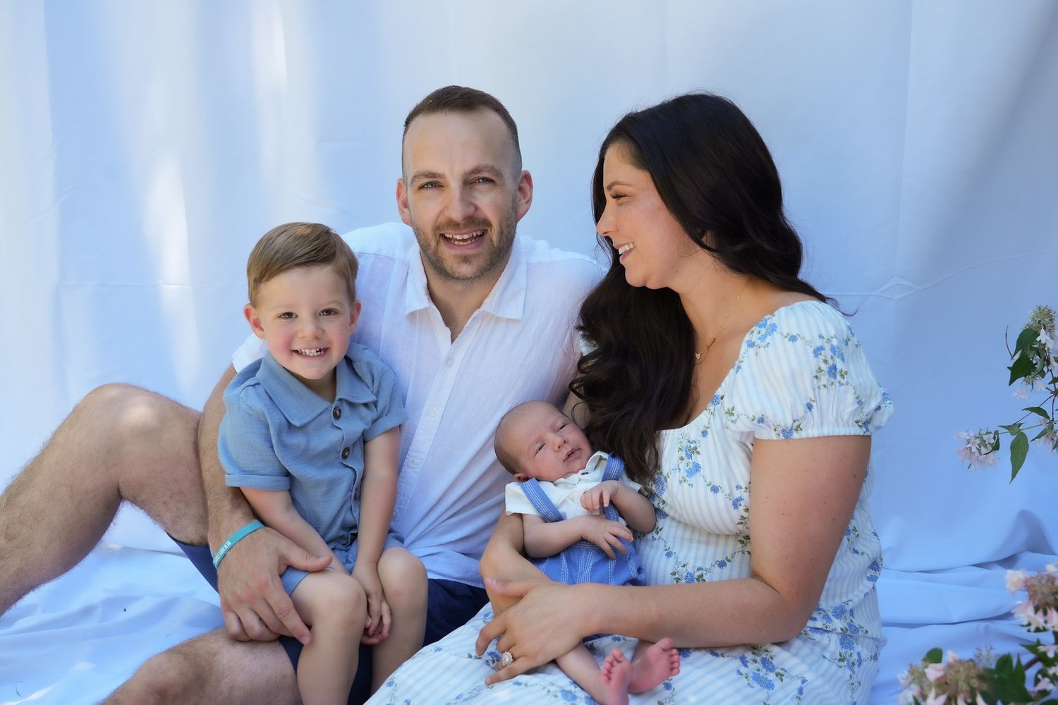 Happy family of four with newborn baby posing together against soft blue backdrop.