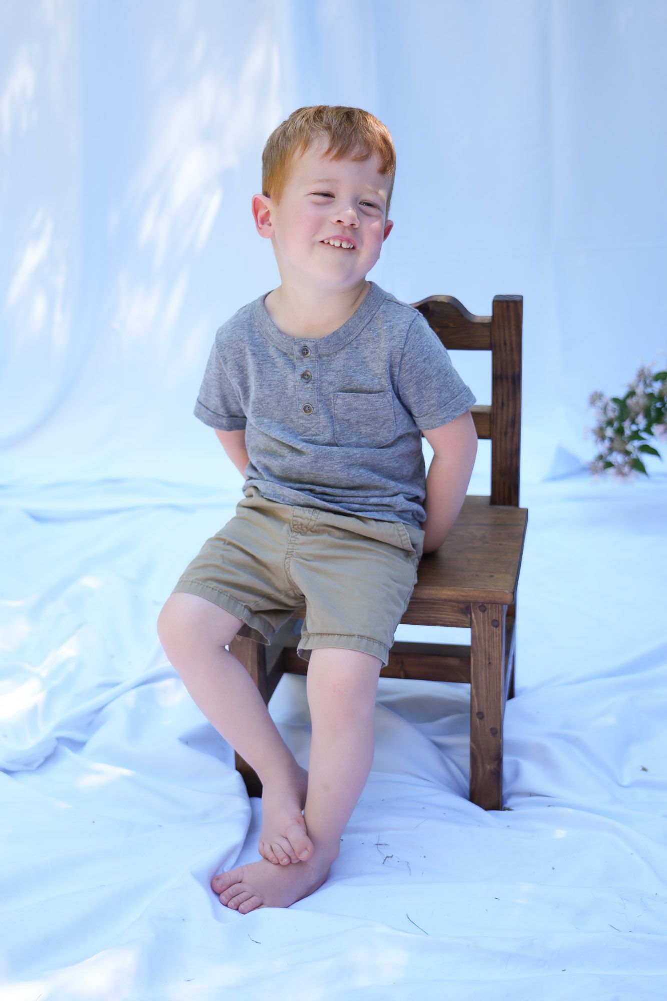 Cute red-haired boy in gray shirt and khaki shorts sitting on wooden chair smiling.