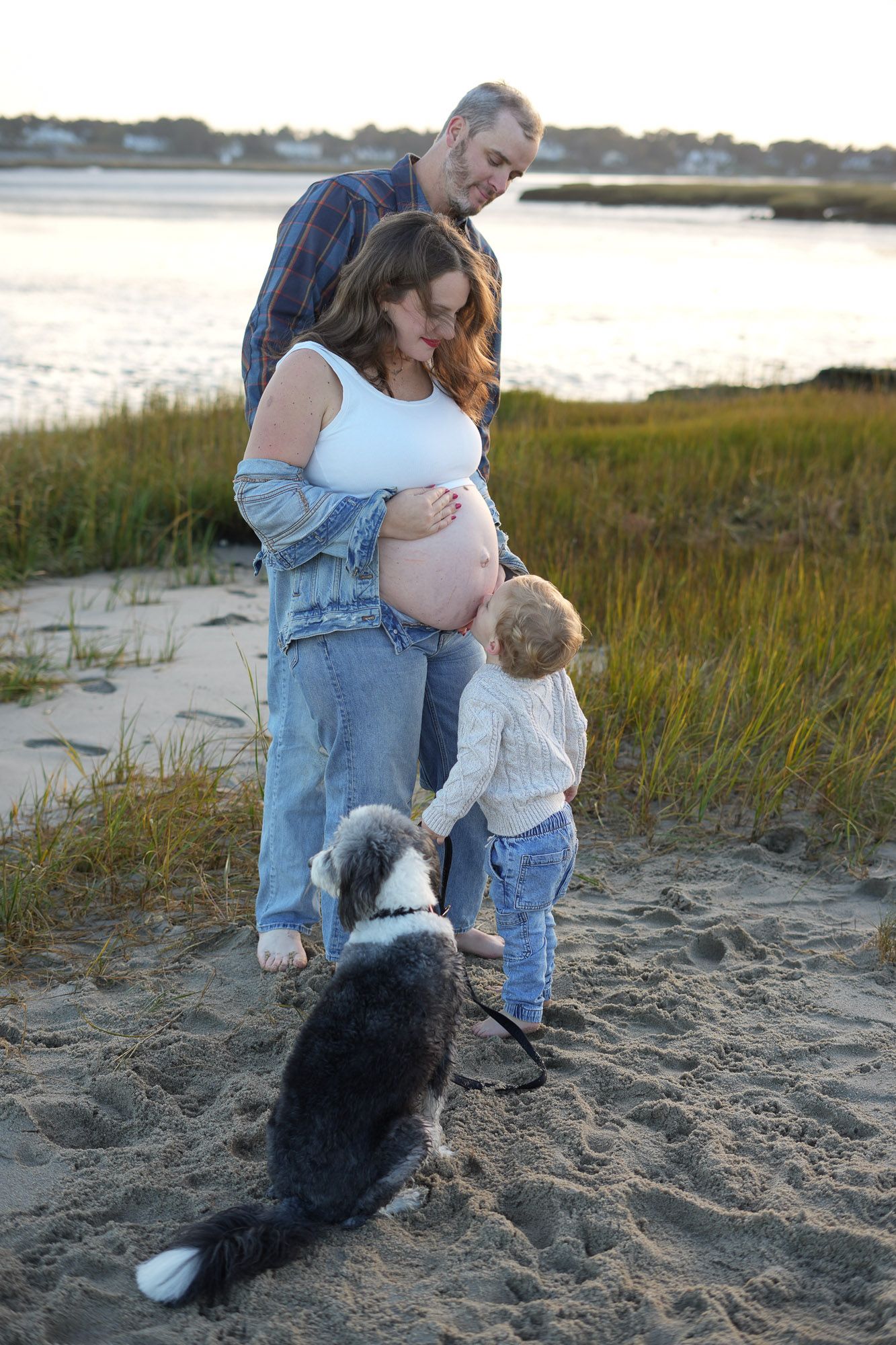 Pregnant couple with toddler touching baby bump and dog on leash on beach at dusk.