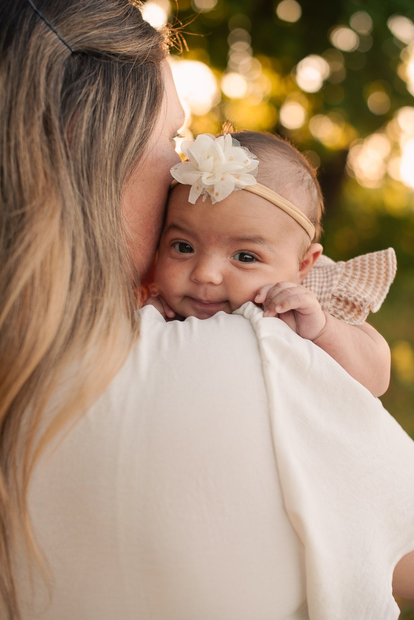 A smiling baby girl with a white flower headband rests on her mother's shoulder outdoors at golden hour.