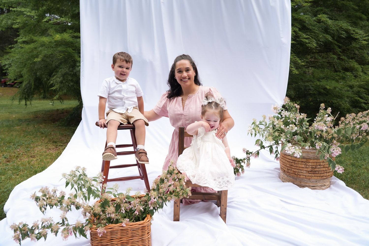 Mother in pink dress smiling with toddler boy on stool and baby girl at outdoor photo session.