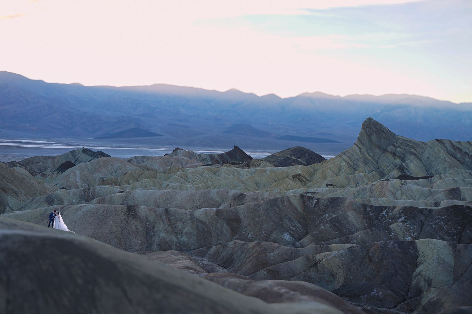 Lone bride and groom in white dress posing among vast rugged Death Valley rock formations at dusk.