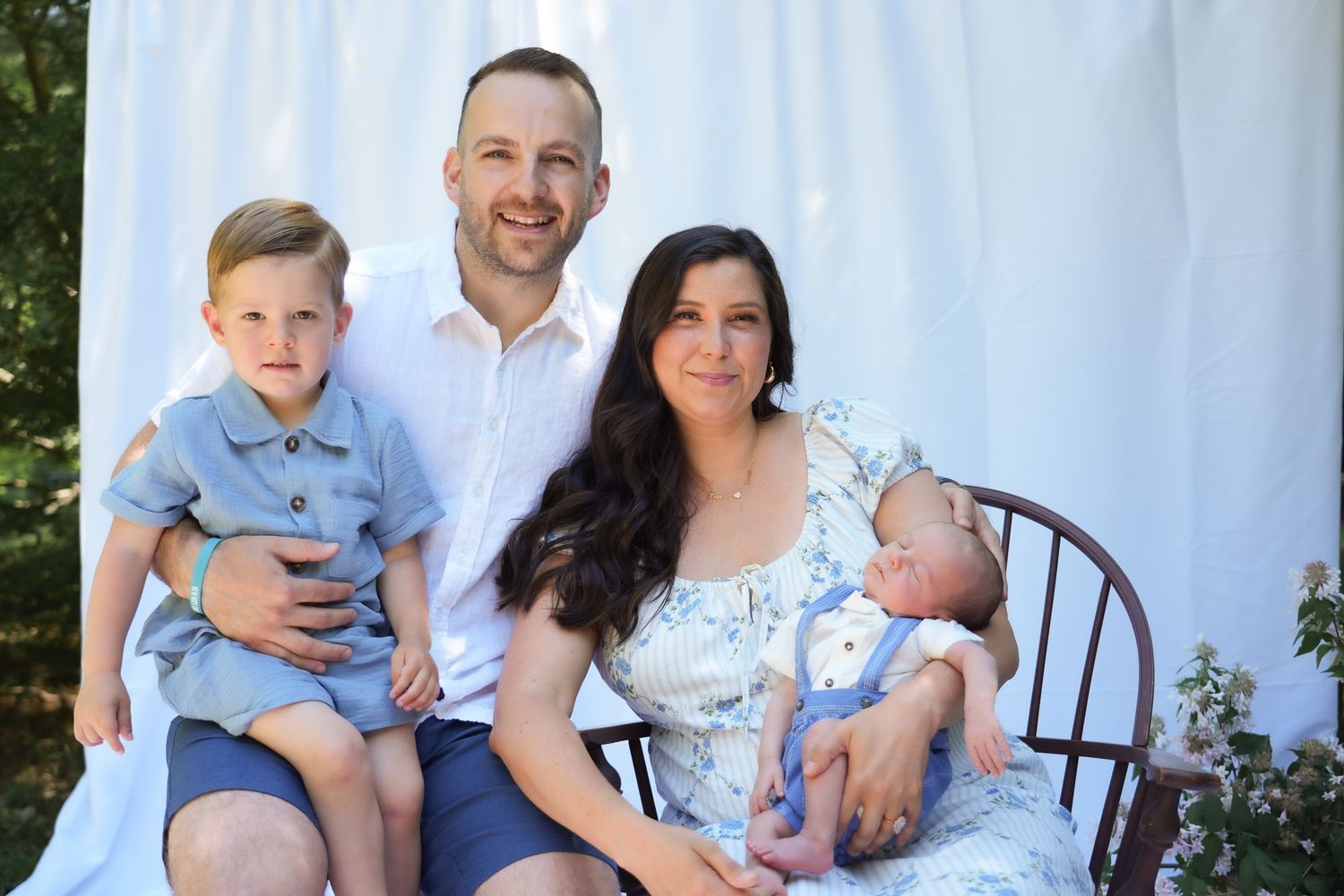 Family portrait with parents, toddler boy, and newborn baby outdoors on white backdrop.