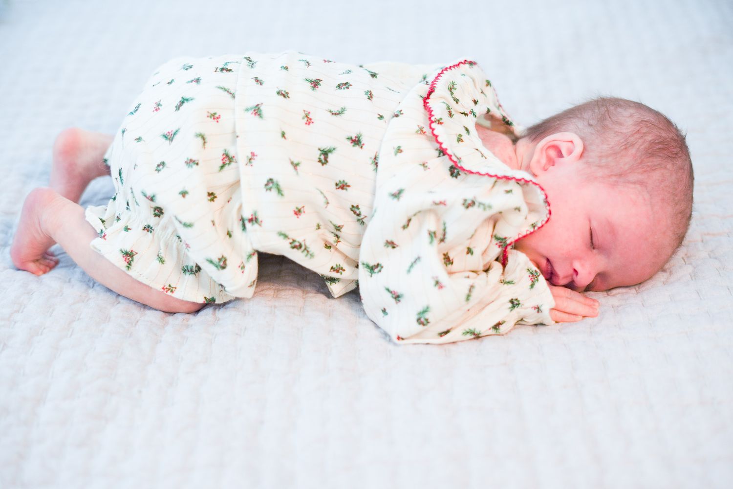 Tiny newborn baby sleeping peacefully in white floral Christmas onesie on soft white blanket.