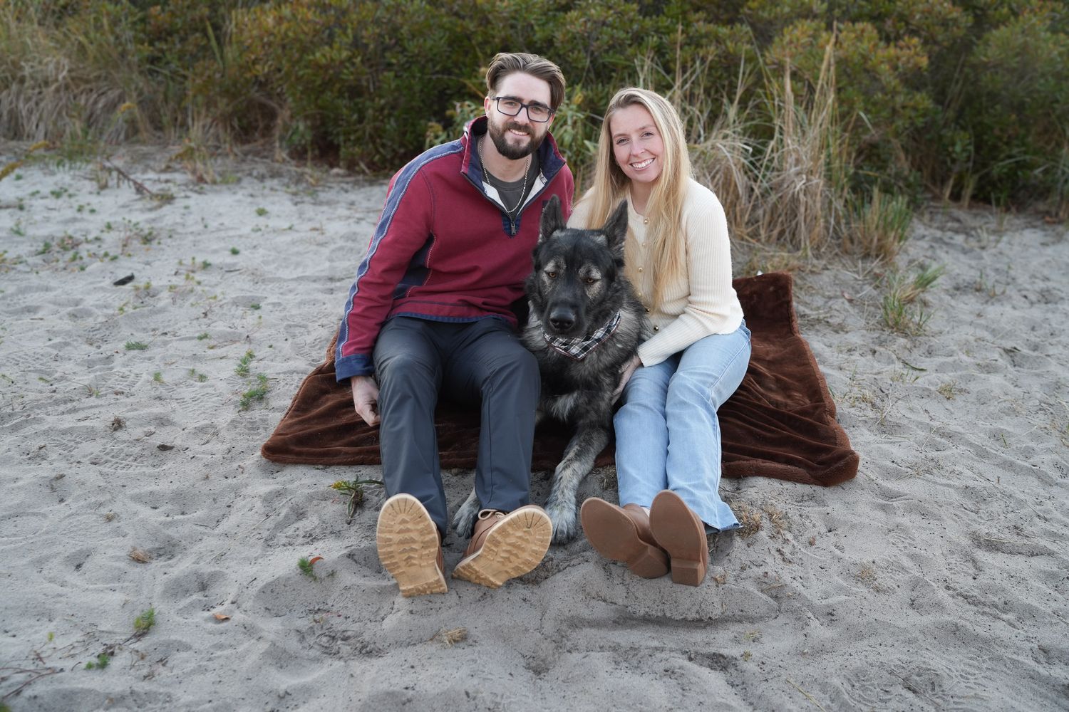 Couple sitting on a blanket in the sand at the beach with their German Shepherd dog between them.