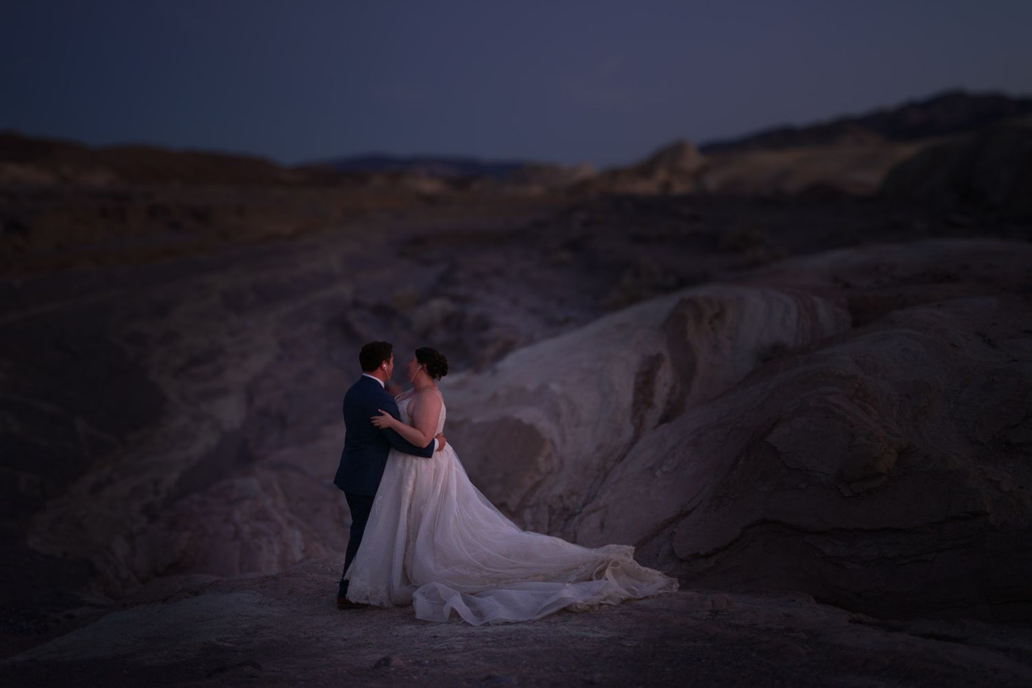 Bride in flowing gown and groom embracing under a twilight sky in a desert landscape.