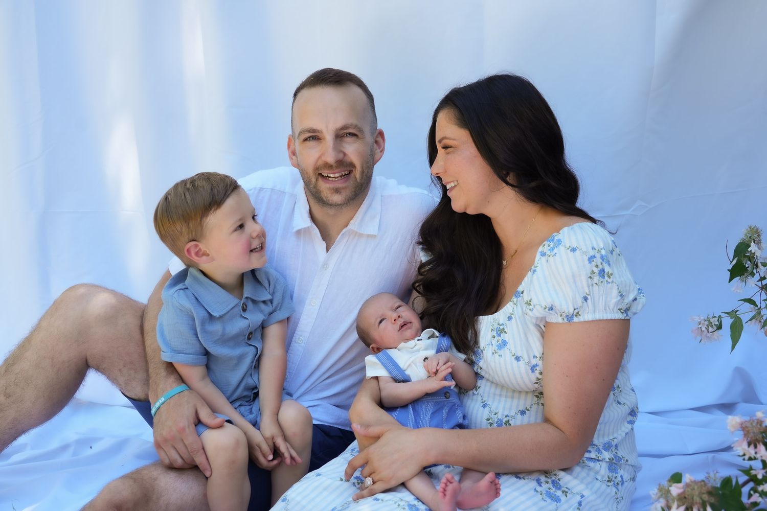 Smiling family of four with newborn baby and toddler son sitting on white fabric.