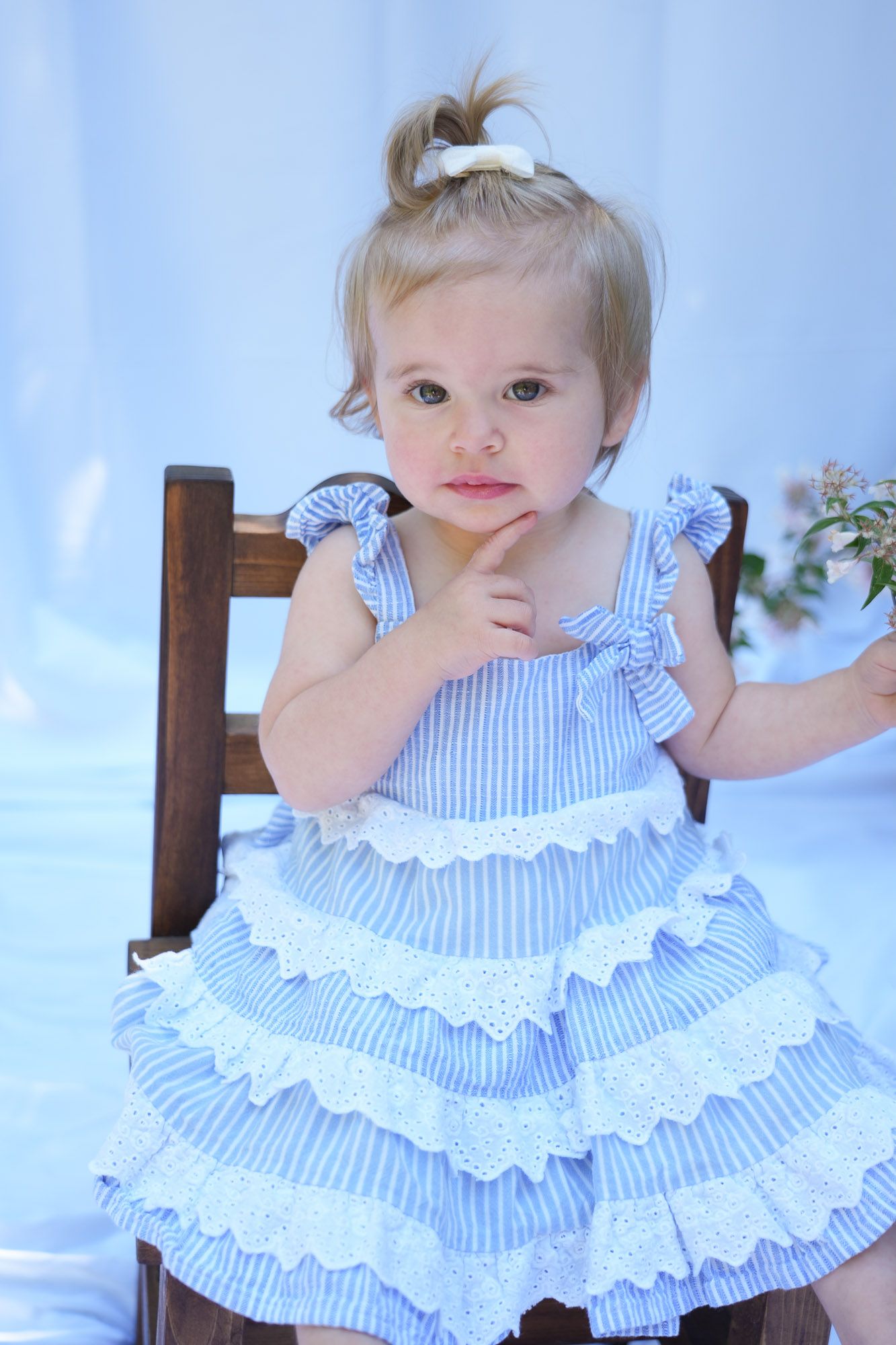 Adorable blonde toddler girl in blue ruffled dress sitting on wooden chair with finger on chin.