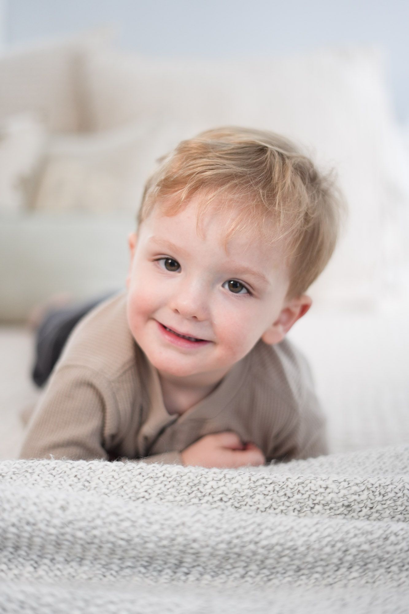 Adorable smiling toddler with blonde hair lying on a cozy gray knitted blanket.