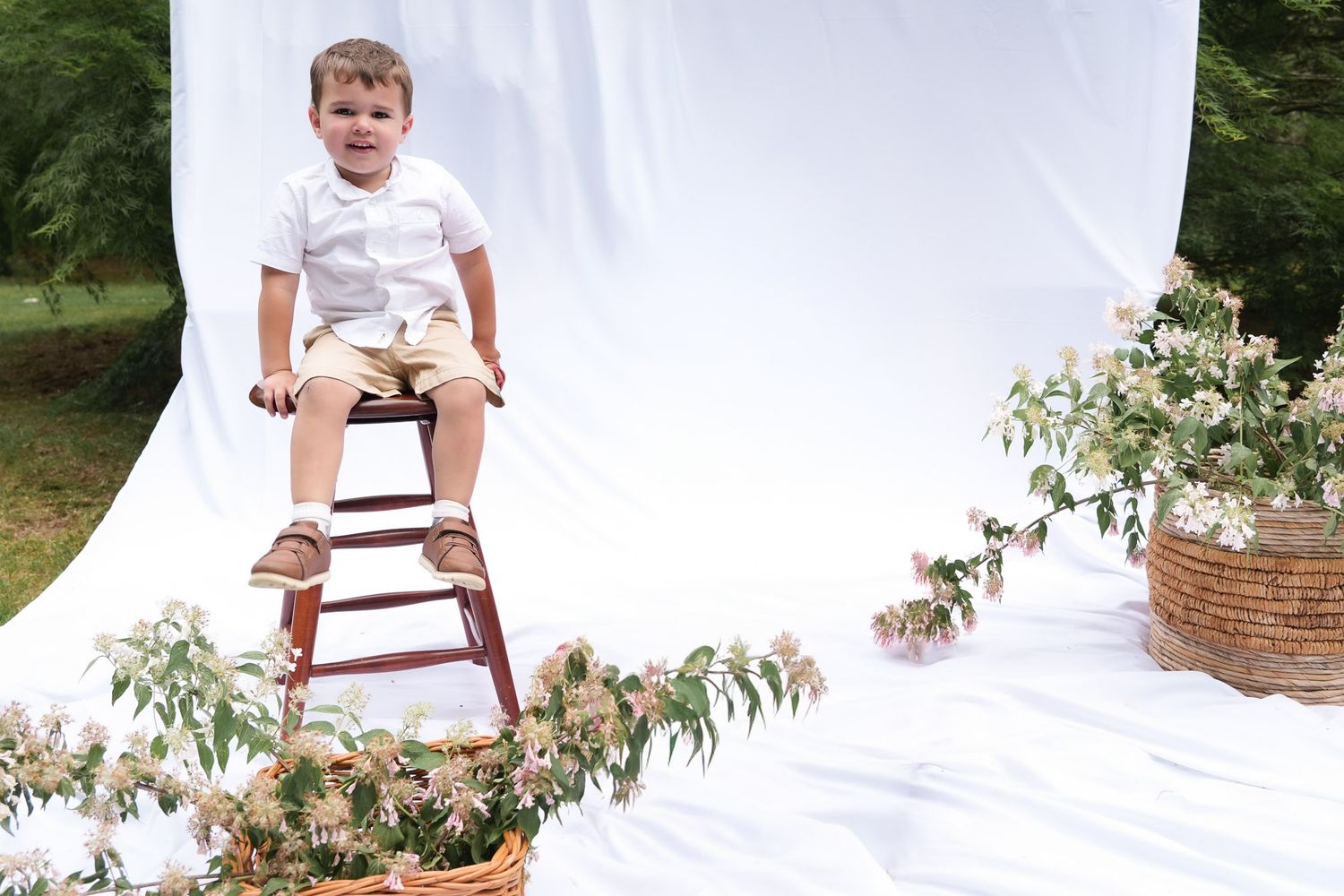 Young boy in white shirt sitting on wooden stool surrounded by flowers at outdoor photo shoot.
