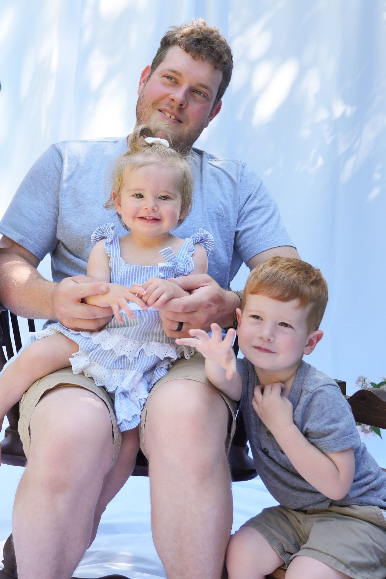 Father sits outdoors holding blonde toddler girl while red-haired young son poses beside them for portrait.