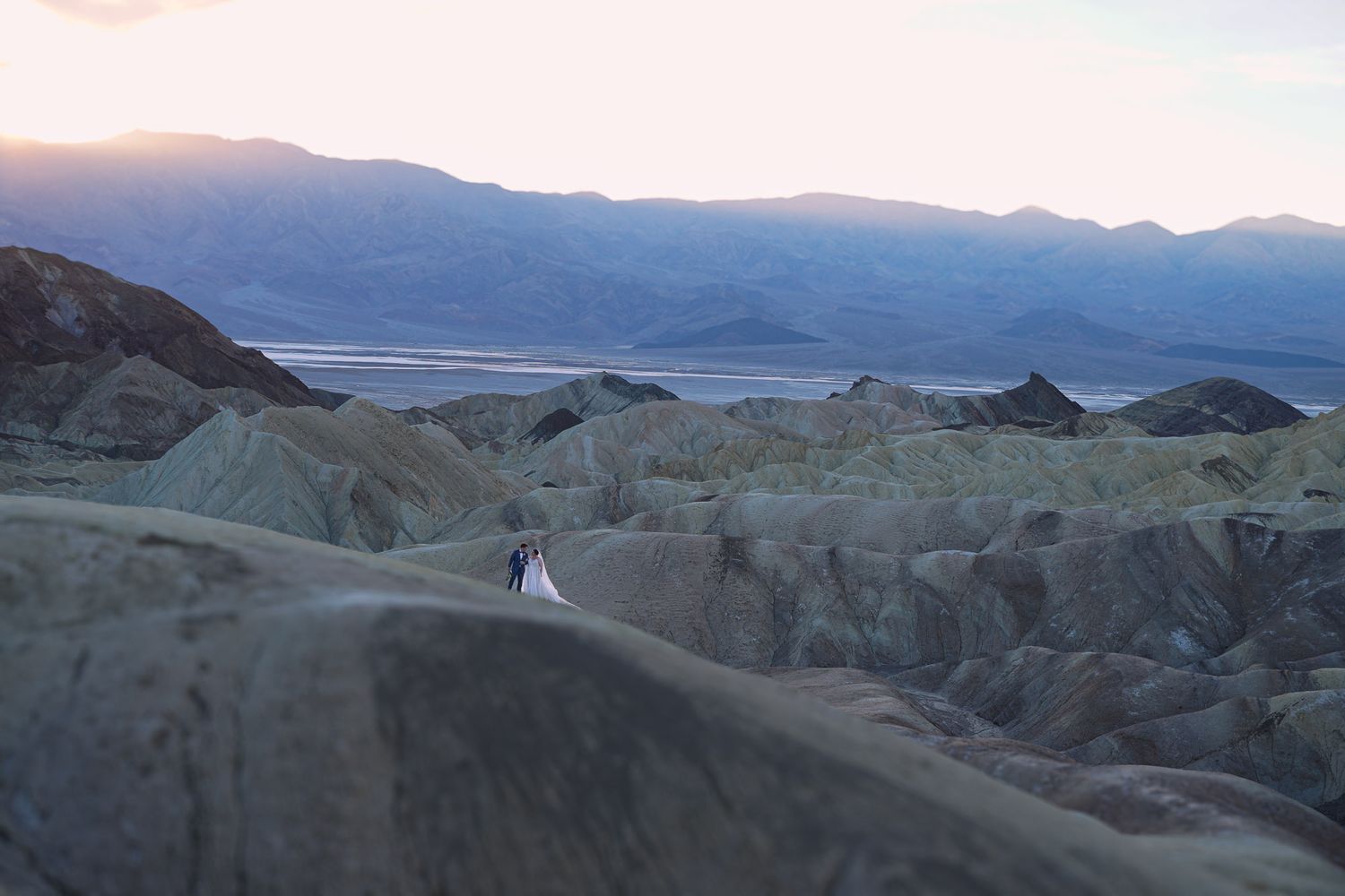 Wedding couple in white attire standing amid stunning eroded rock formations at Death Valley at sunset.