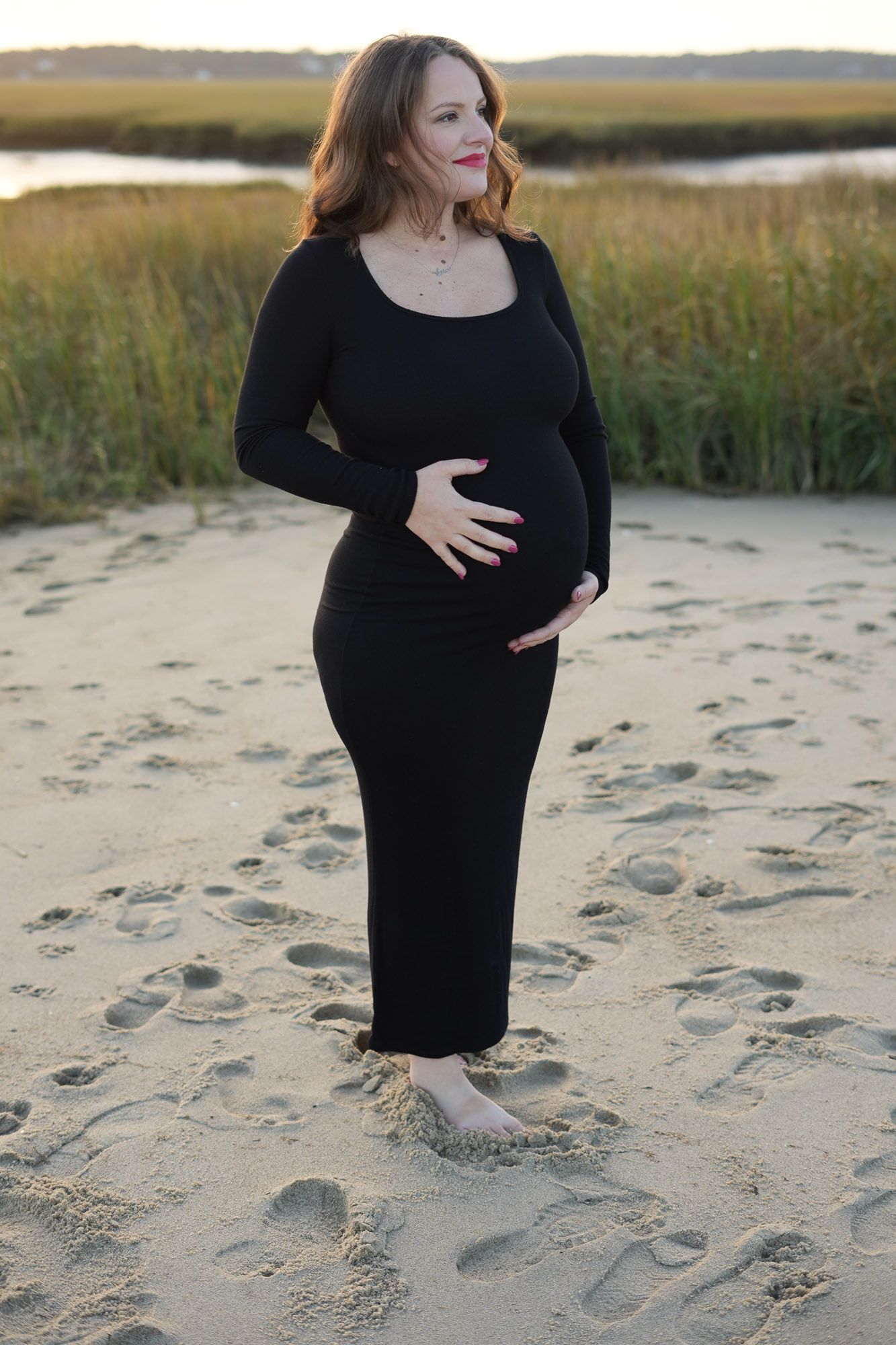 Pregnant woman in black maxi dress cradling baby bump while standing barefoot on sandy beach.
